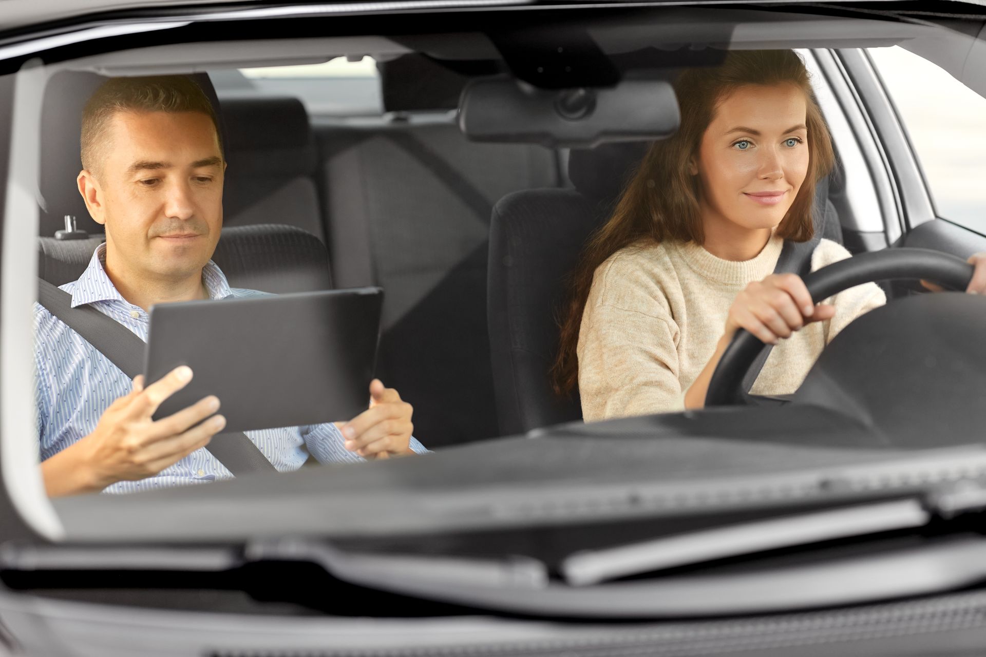 Driving instructor with tablet guides student driver during behind-the-wheel lesson.