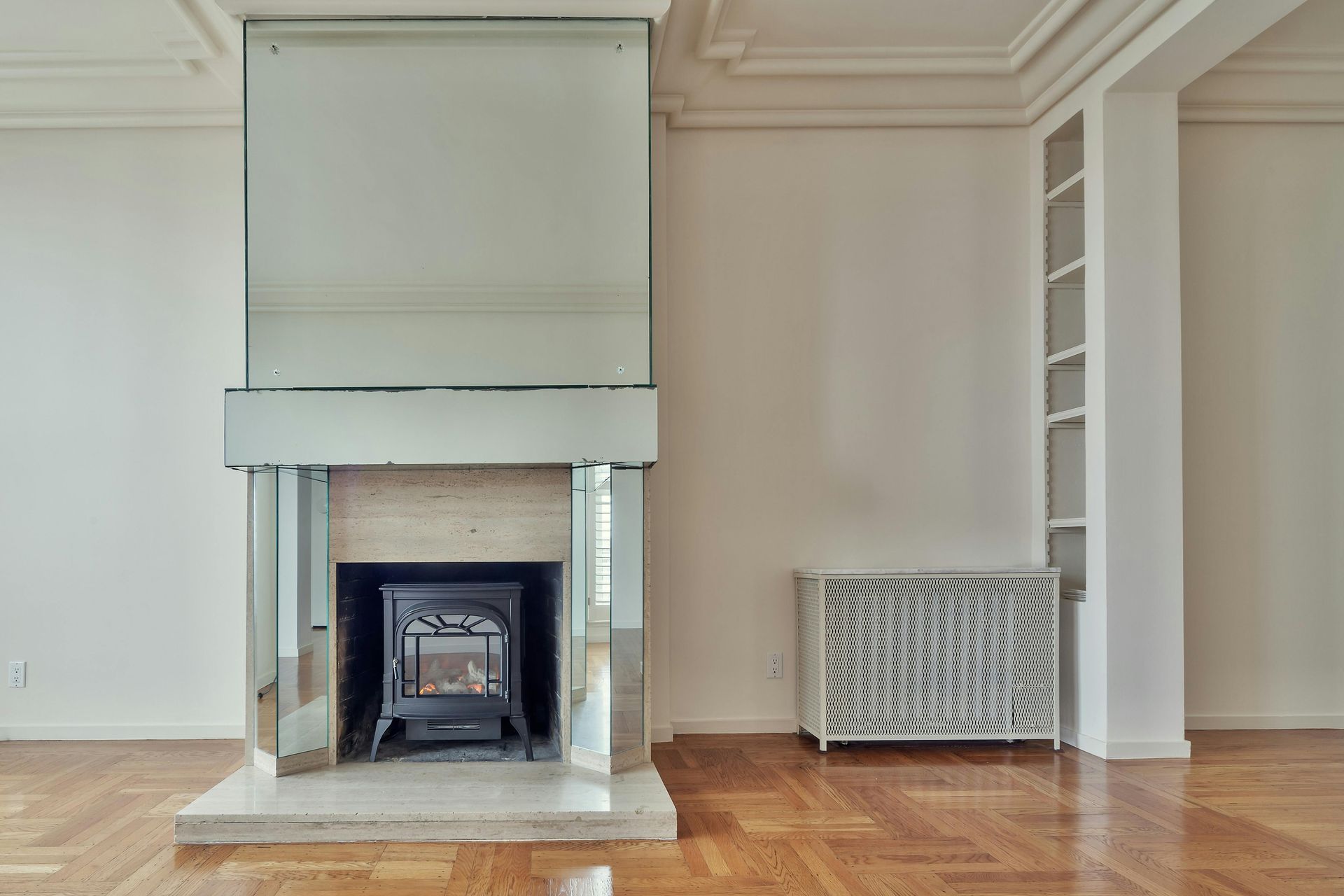 Empty room with a mirror-covered fireplace, built-in shelves, and parquet flooring.