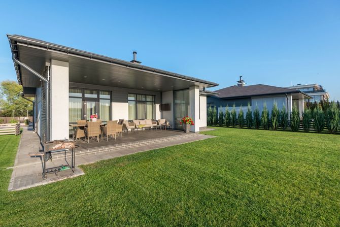 Modern house with a covered patio, outdoor seating, and a large green lawn under a blue sky