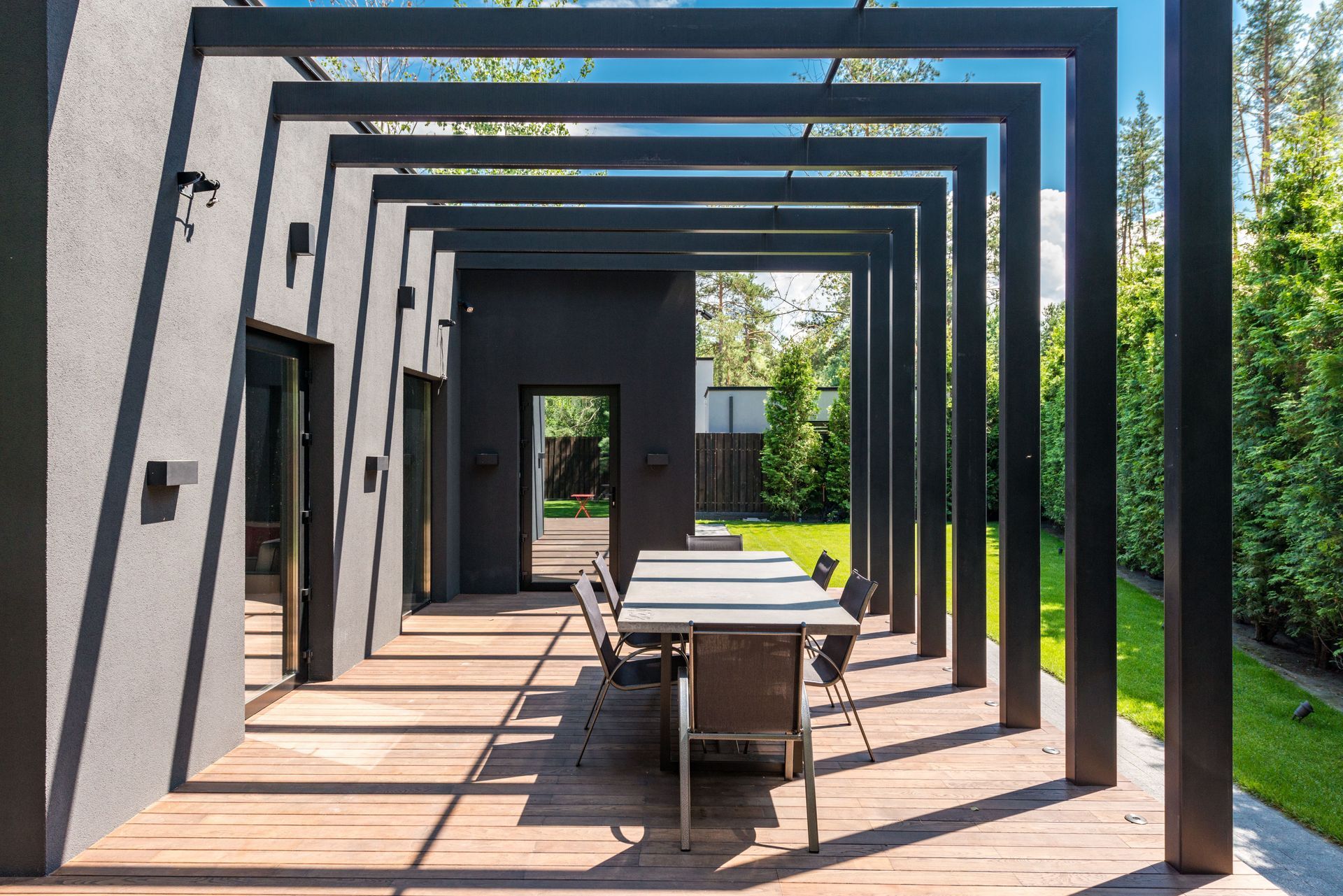 Modern patio with black pergola frames, a long table, and chairs in bright sunlight.