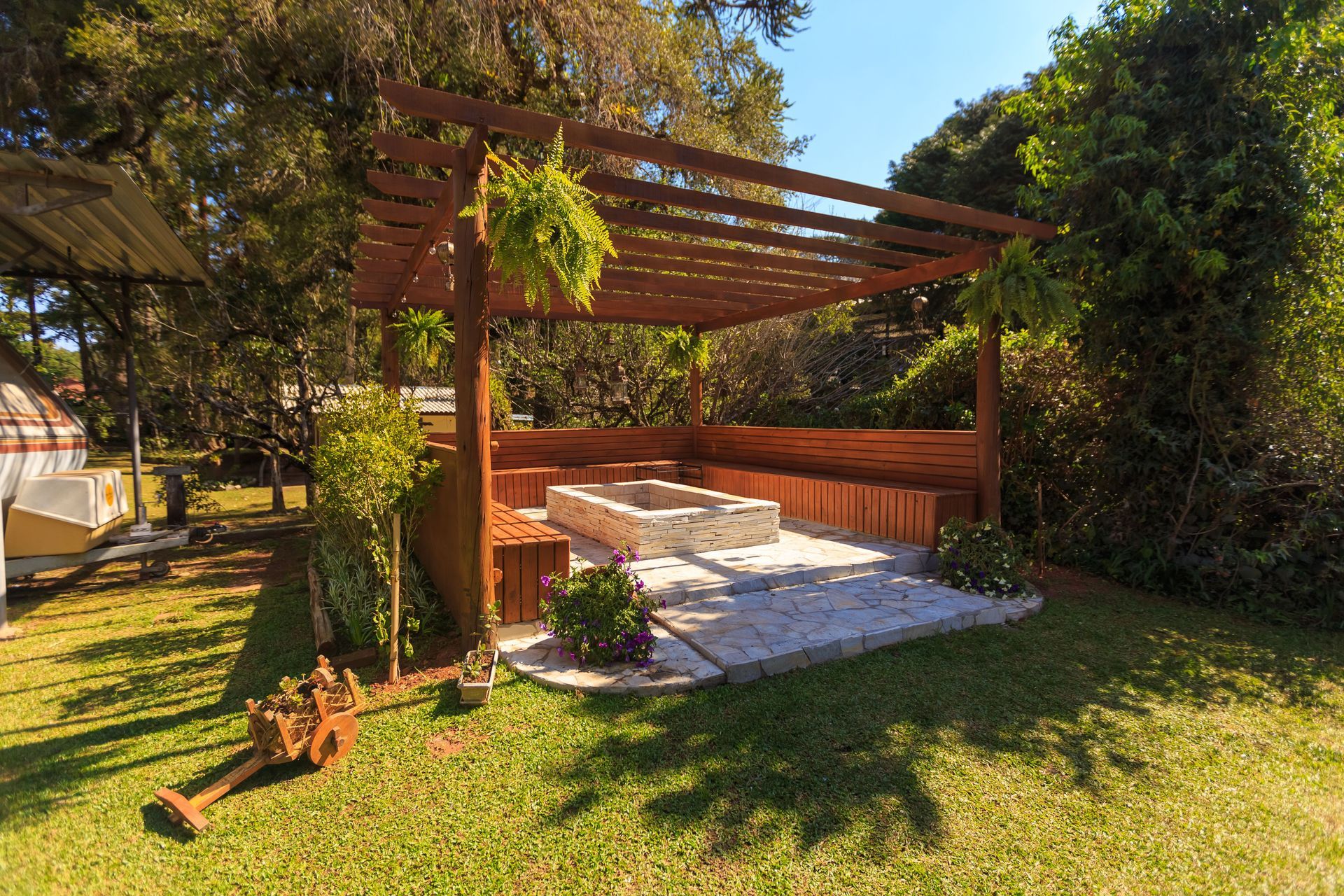 Wooden pergola over a stone patio in a sunny backyard, surrounded by grass and trees.