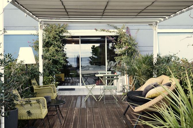 Outdoor patio with wooden deck, lounge chairs, small table, plants, and sliding glass doors under a roofed pergola