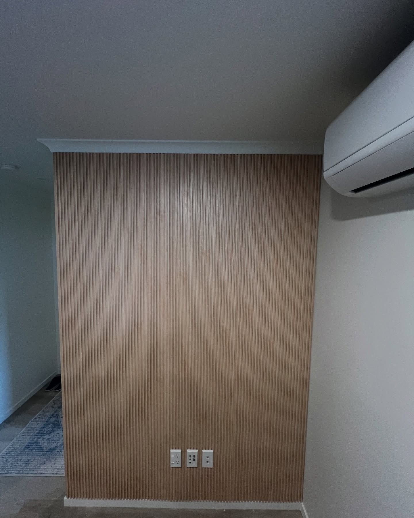 A wall with a textured, vertical wooden panel design, three electrical outlets, and an air conditioner.