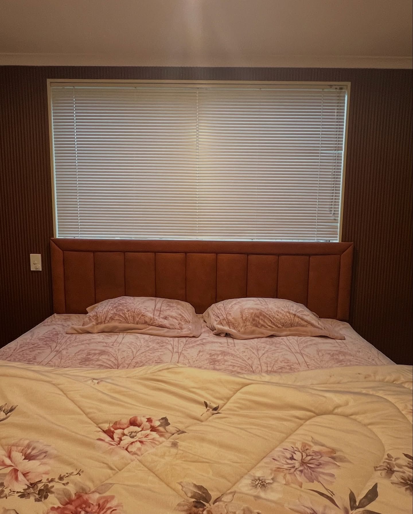 Bed with headboard, two pillows, floral comforter, and a window with blinds.