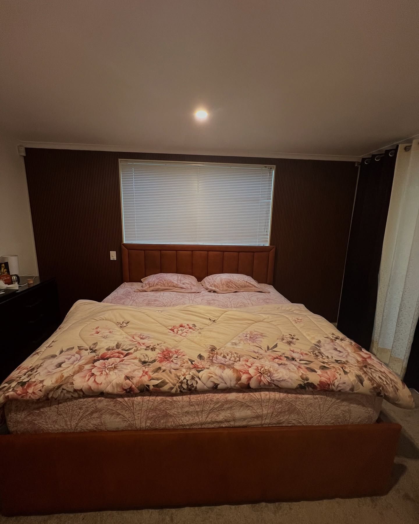 Brown bed with floral comforter in a bedroom with blinds and dark brown walls.