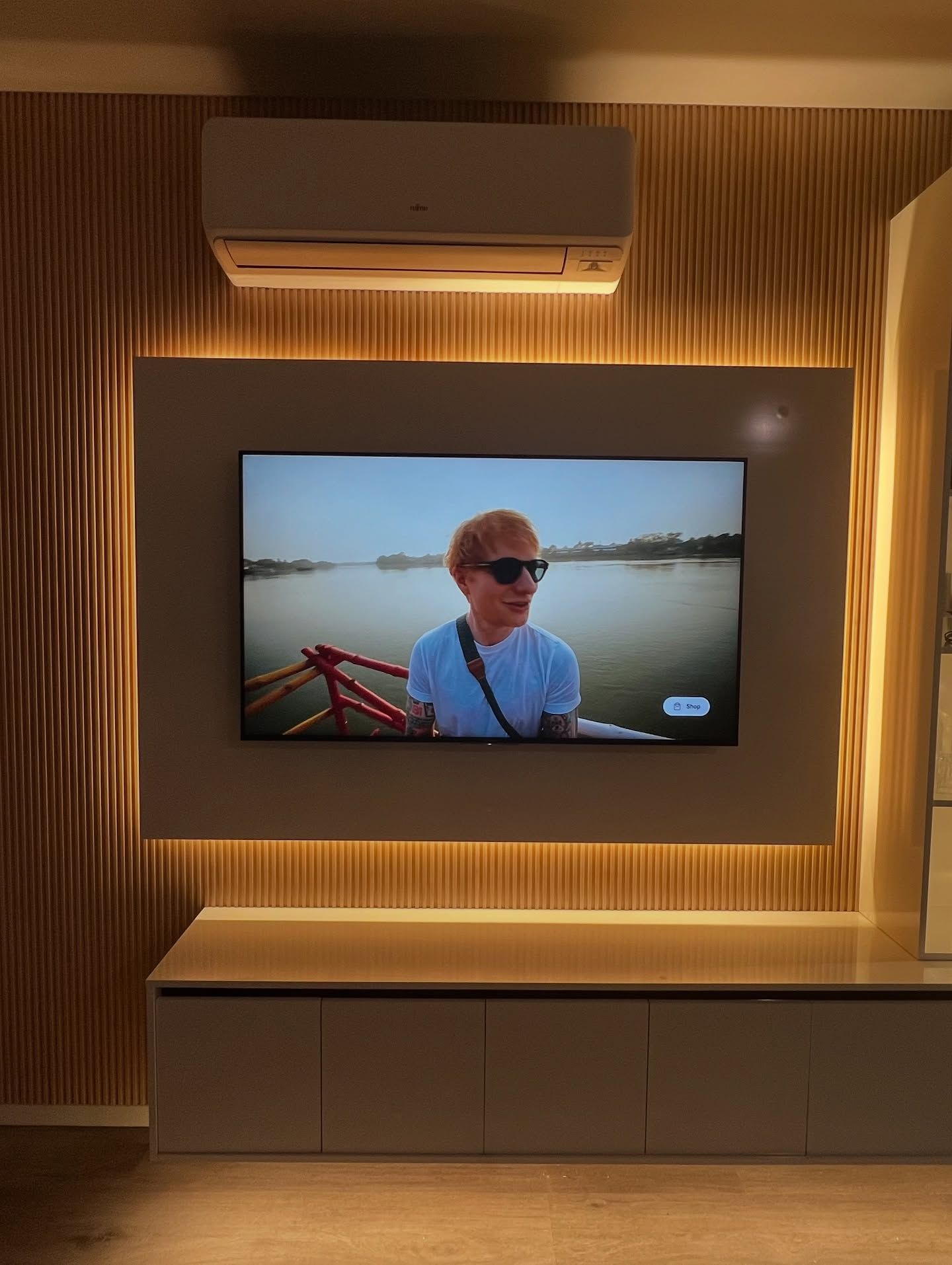 TV mounted on a wall with wood paneling, air conditioner above. Person on screen. Cabinet below.