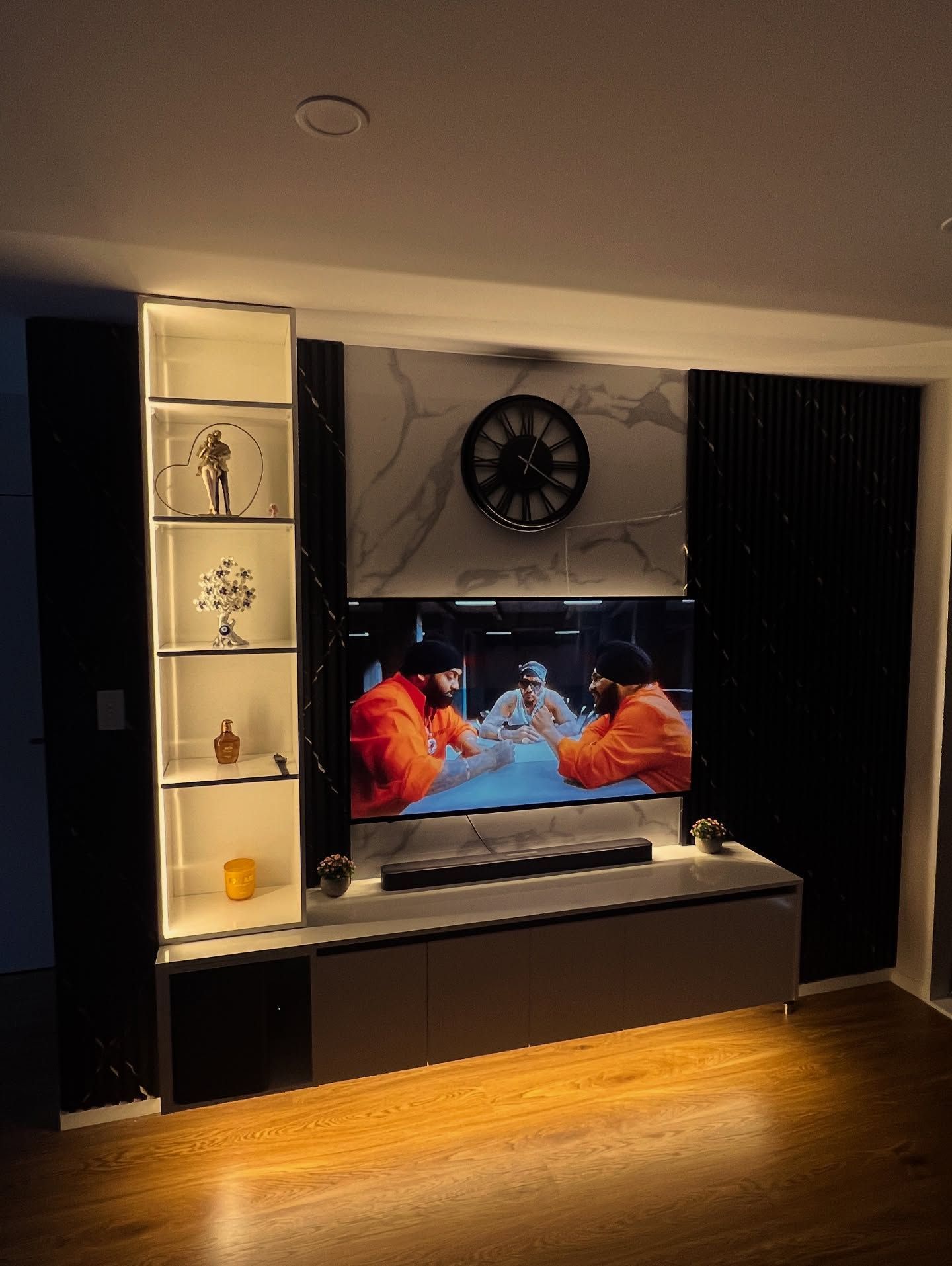Living room entertainment center with TV, shelves, clock, and warm lighting.