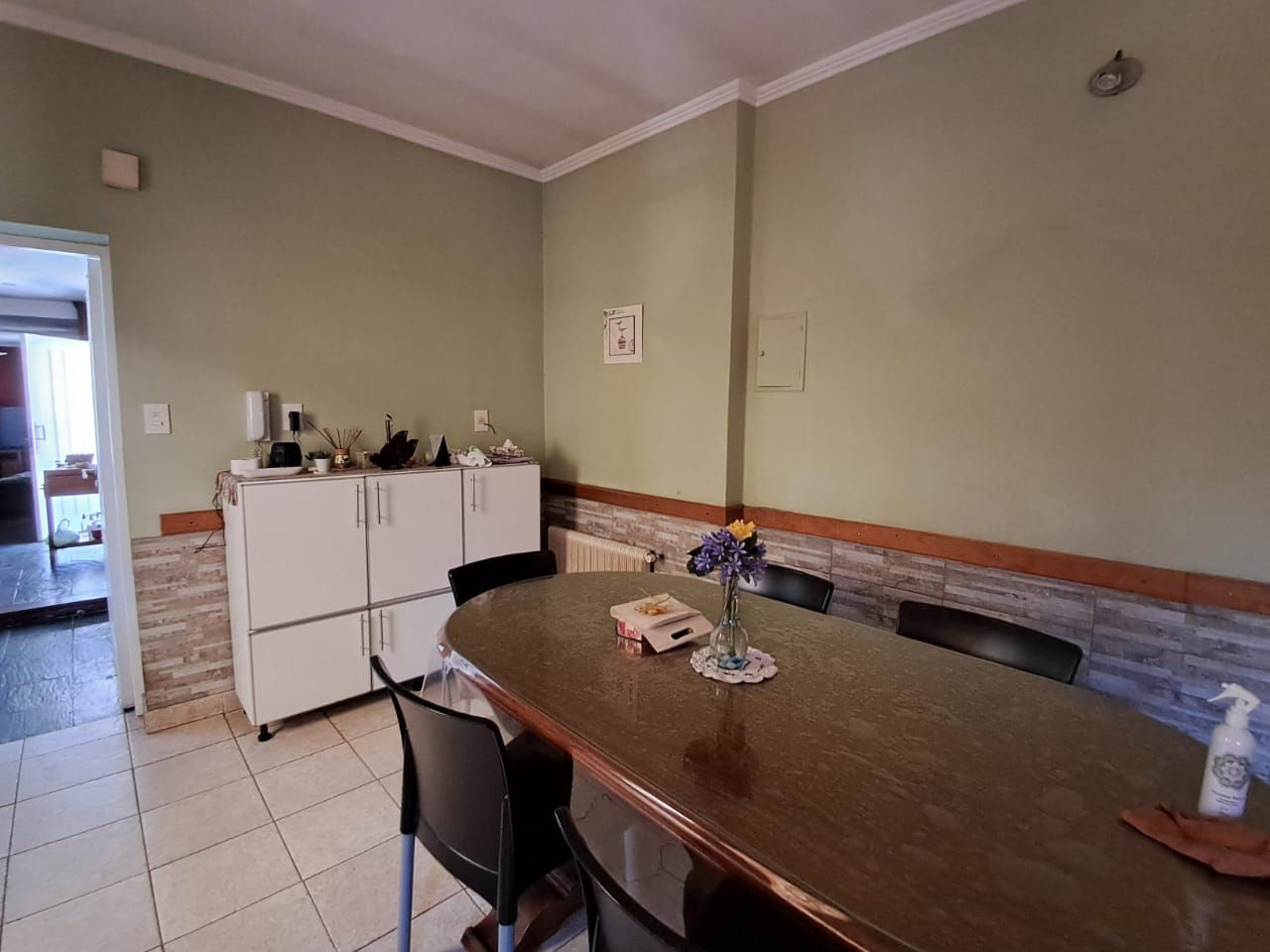 Comedor con mesa ovalada, sillas, armarios, paredes verde salvia y suelo de baldosas.