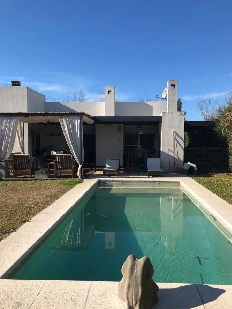 Patio trasero con piscina rectangular frente a una casa blanca con terraza cubierta, cielo azul despejado.