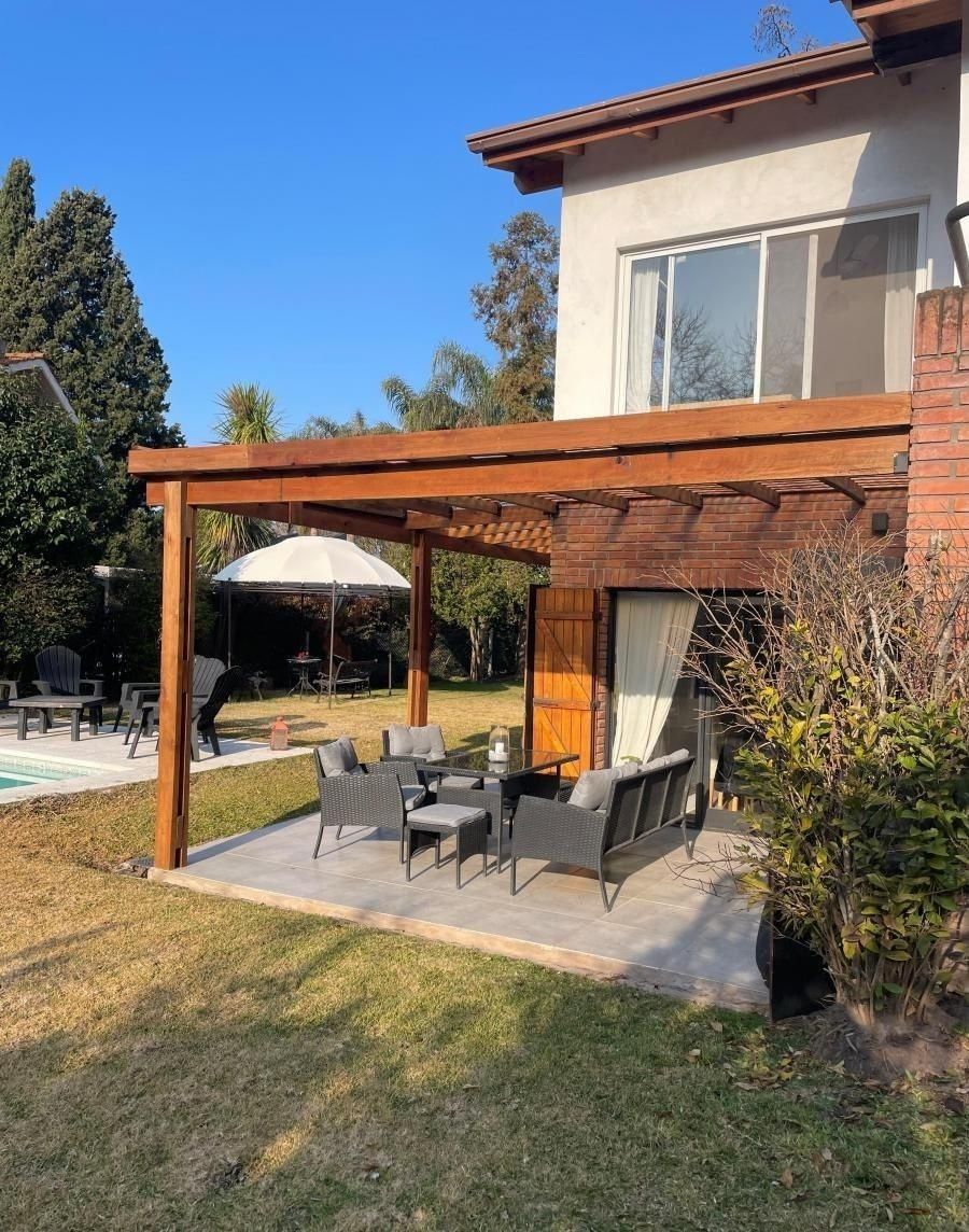 Patio con pérgola de madera, zona de estar y sombrilla, cerca de una casa con ventana.