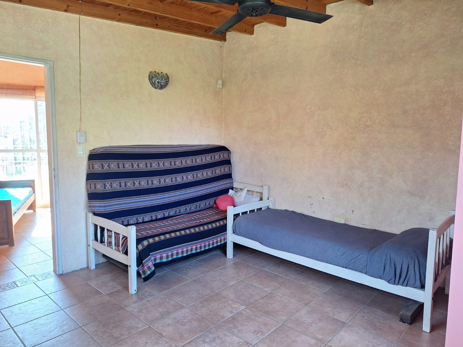 Habitación con futón, cama individual y puerta abierta; paredes de color beige claro y suelo de baldosas.