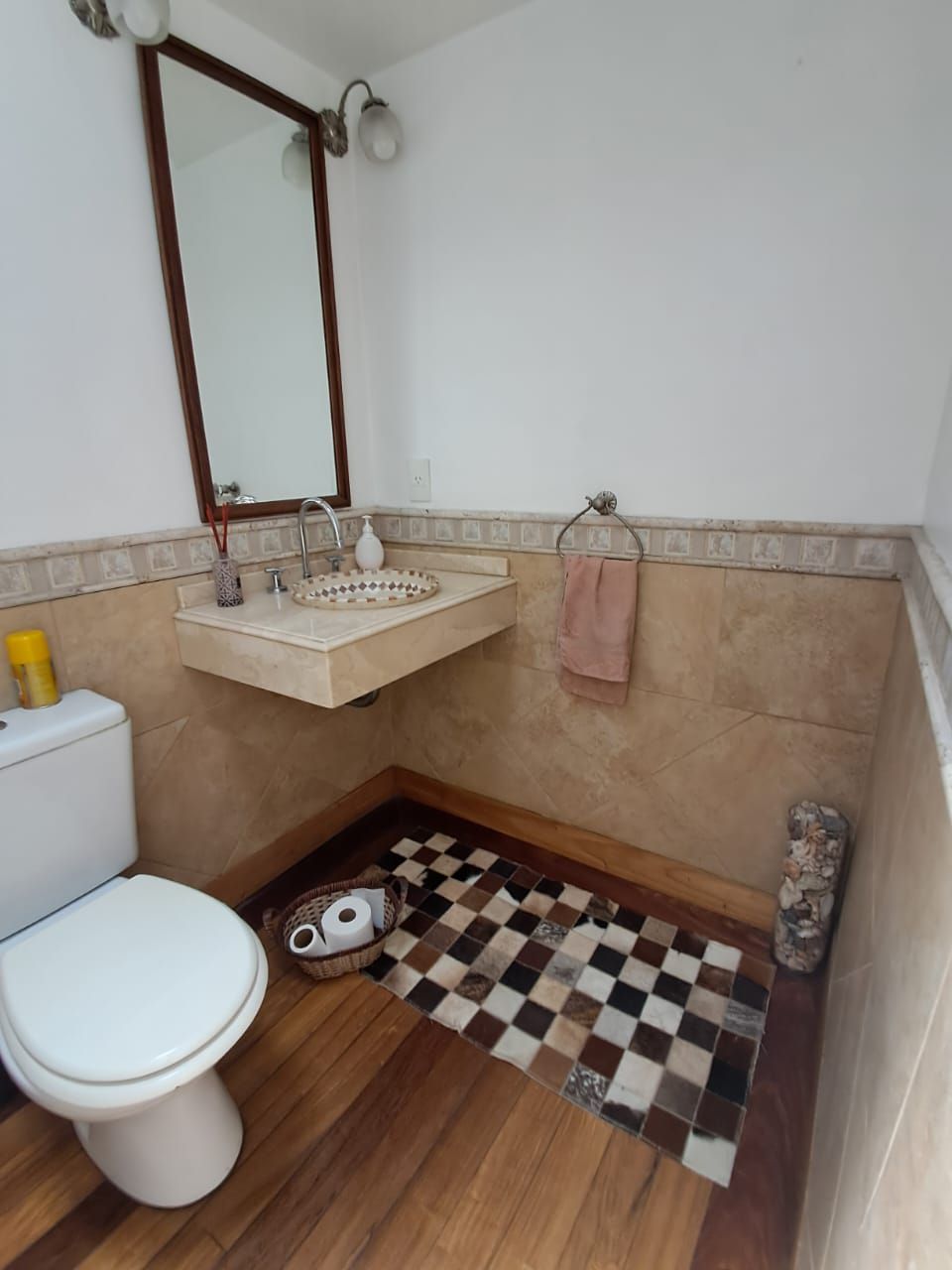 Baño con inodoro, lavabo, espejo y alfombra de cuadros sobre suelo de madera.