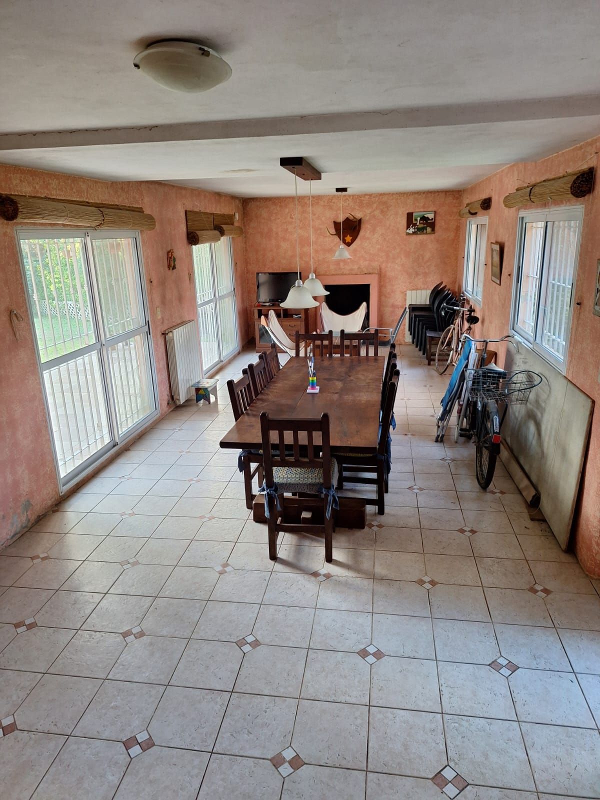 Mesa de comedor larga dispuesta en una habitación con suelo de baldosas, ventanas y chimenea.