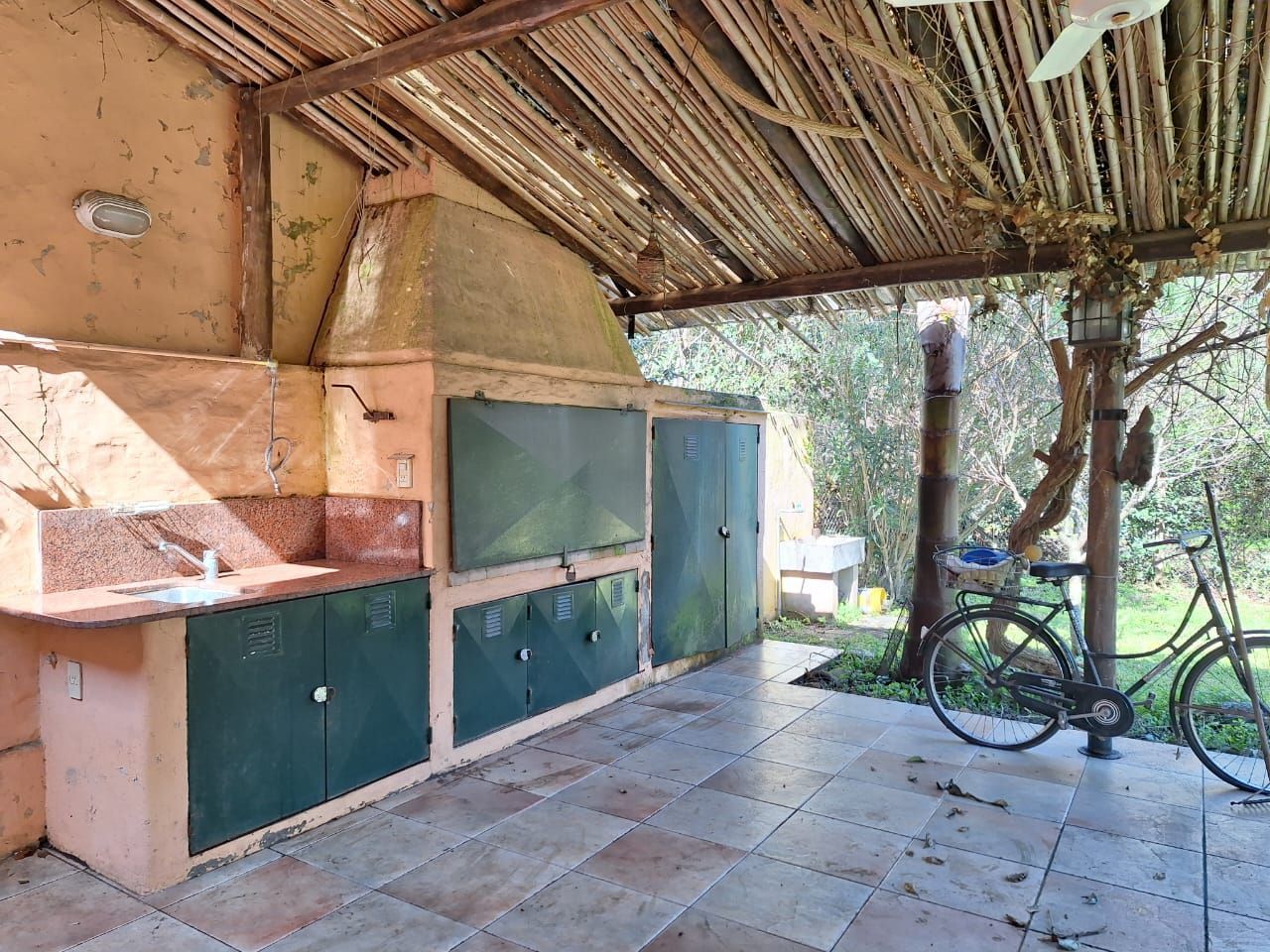 Cocina exterior con parrilla integrada, armarios, fregadero y bicicleta. Techo rústico de madera.