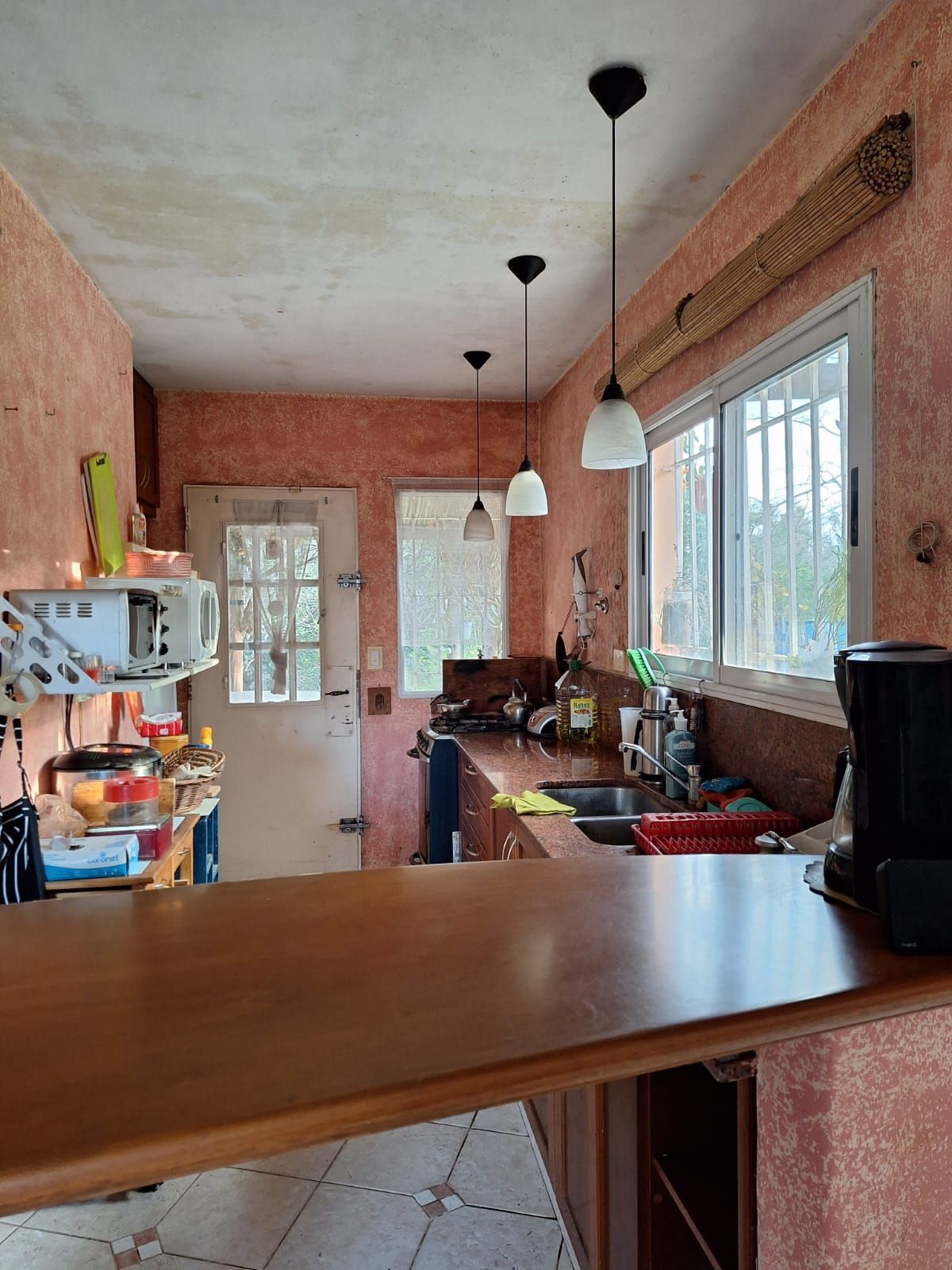 Cocina con barra de madera, fregadero y lámparas colgantes. Paredes rosas y una ventana.