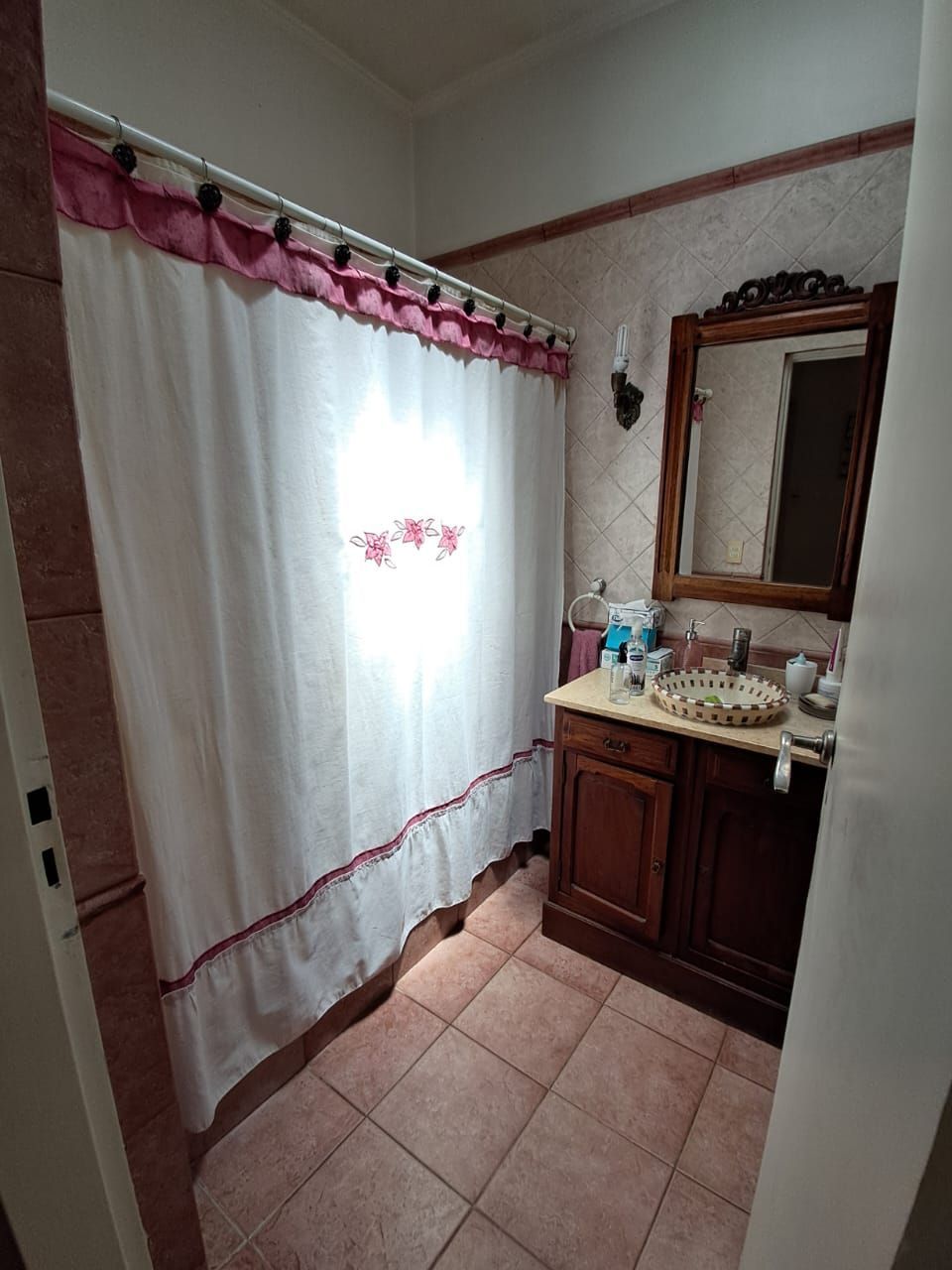 Baño con cortina de ducha, tocador con espejo y suelo de baldosas.