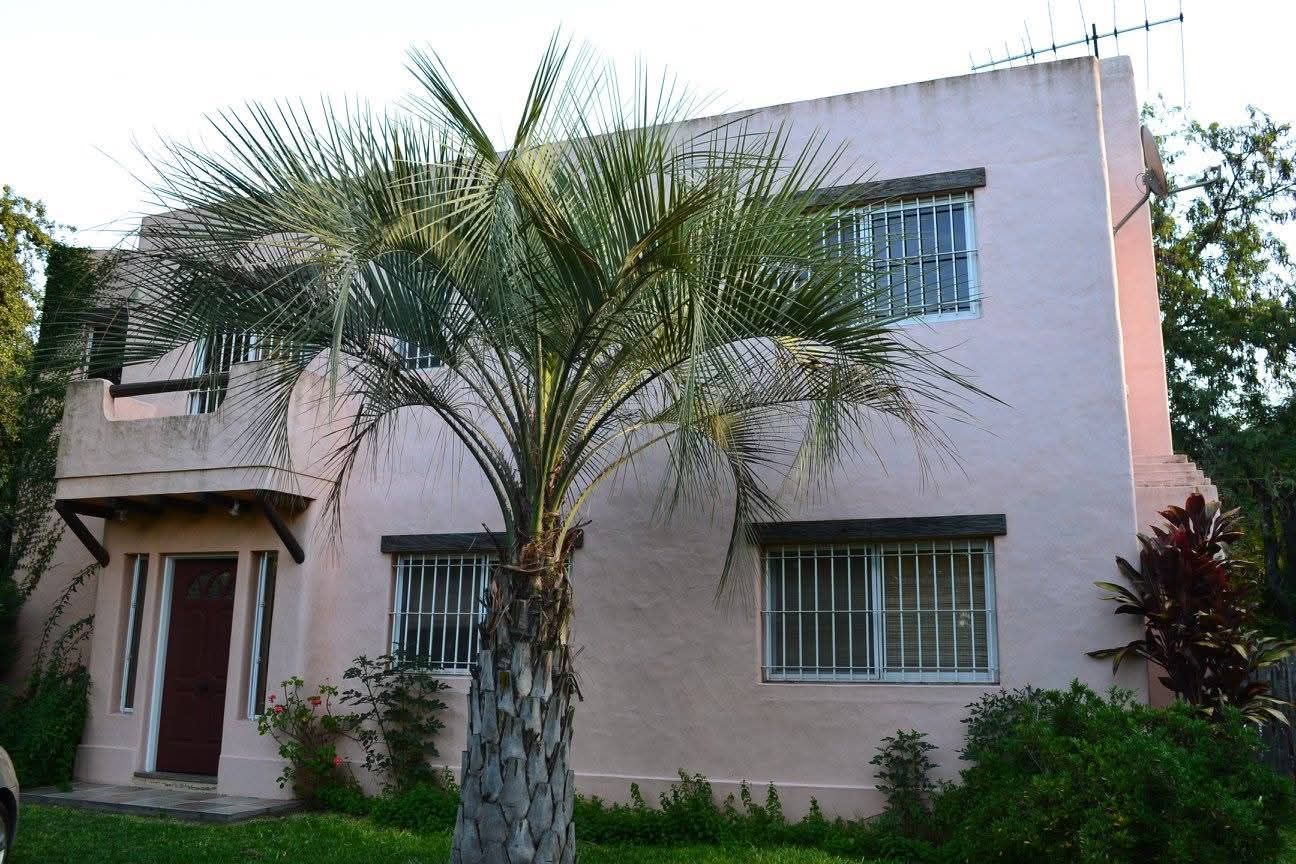Casa rosa de dos plantas con ventanas enrejadas, palmera delante, puerta de madera oscura.