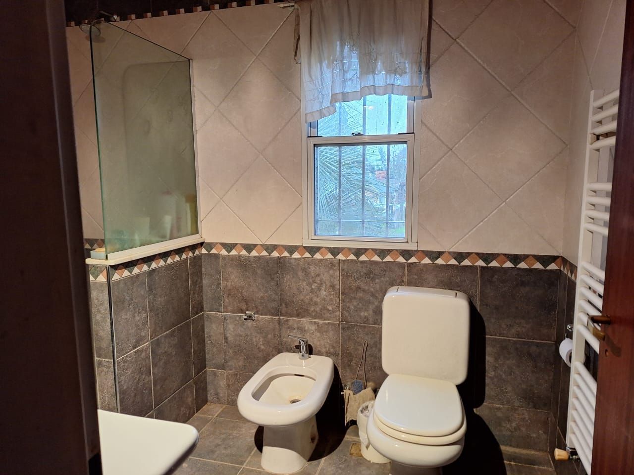 Baño con paredes alicatadas, inodoro, bidé, ducha y ventana con cortina. El toallero está a la derecha.