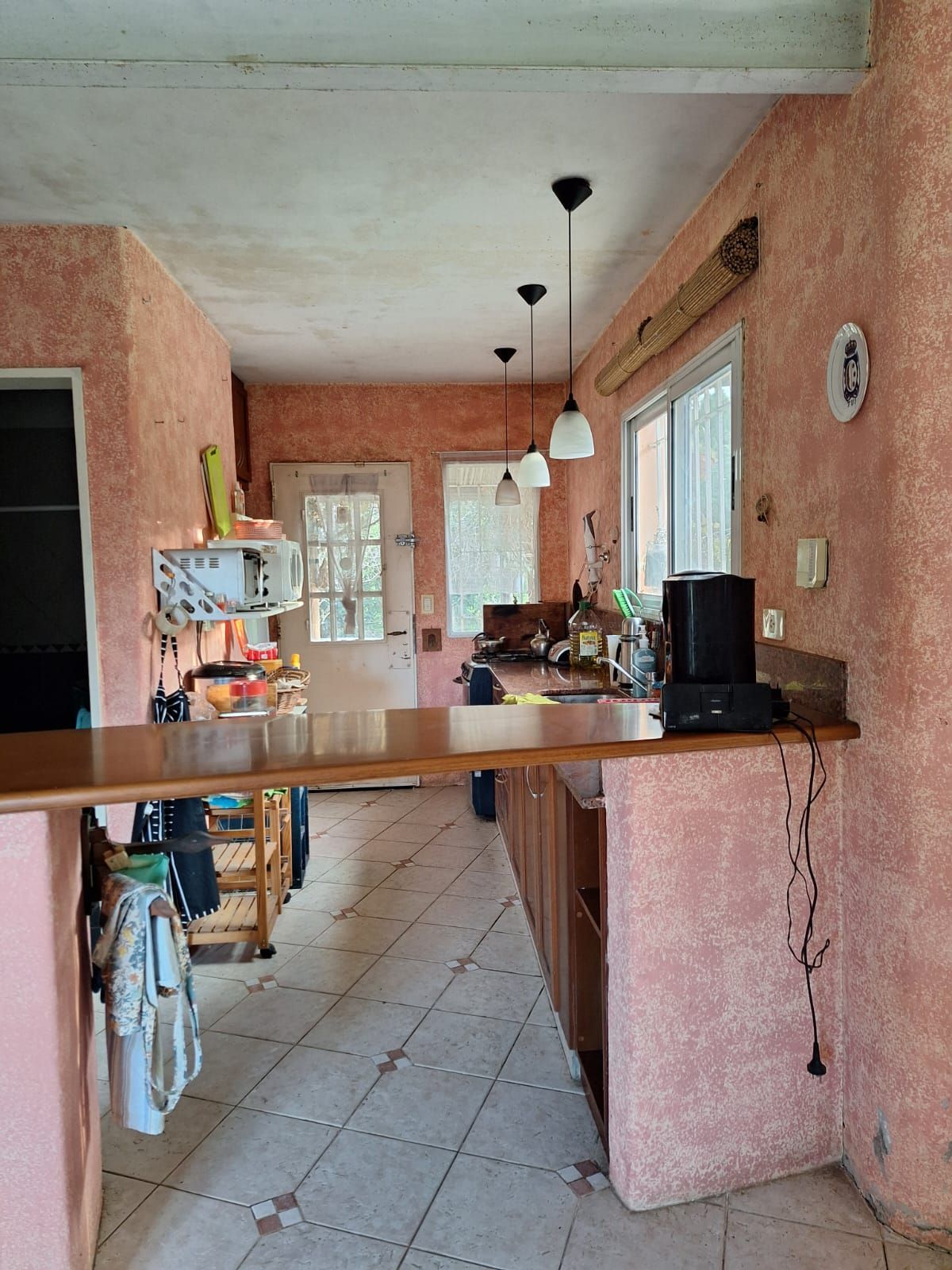 Cocina con paredes texturizadas de color rosa, encimera de madera, suelo de baldosas y tres lámparas colgantes.