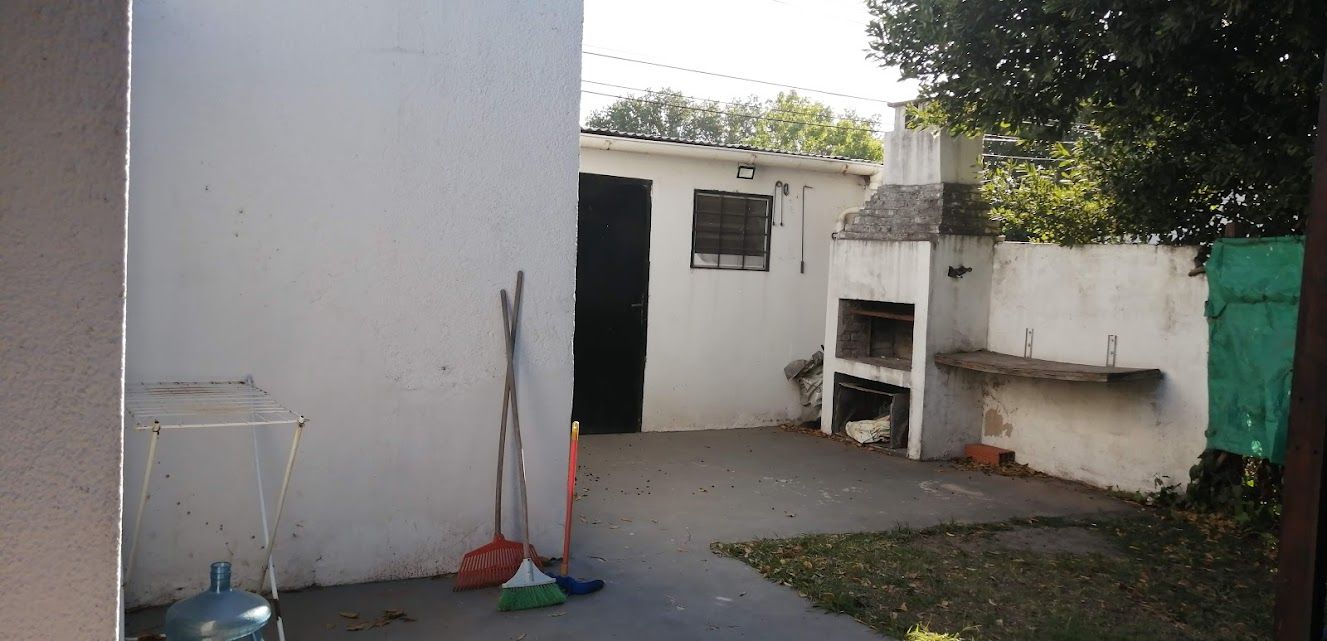 Patio trasero con una pared blanca, una parrilla, un cobertizo y algo de vegetación.