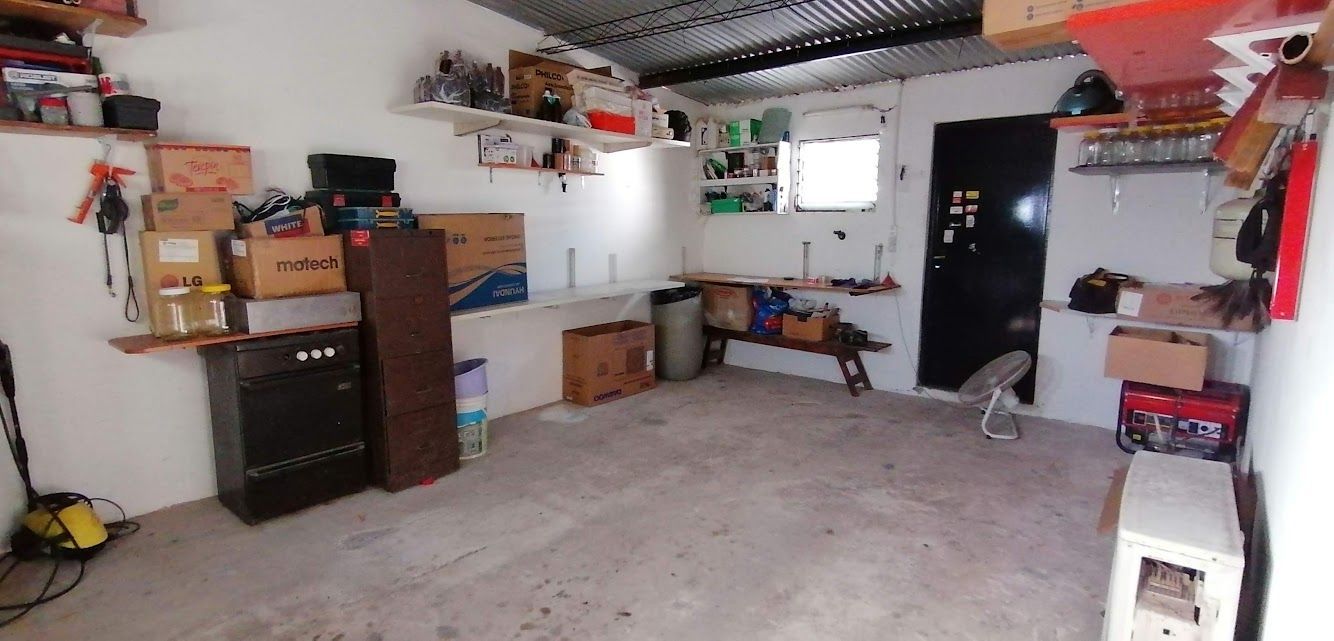 Garaje desordenado con estantes, cajas y una puerta negra. Suelo de hormigón y techo de chapa ondulada.