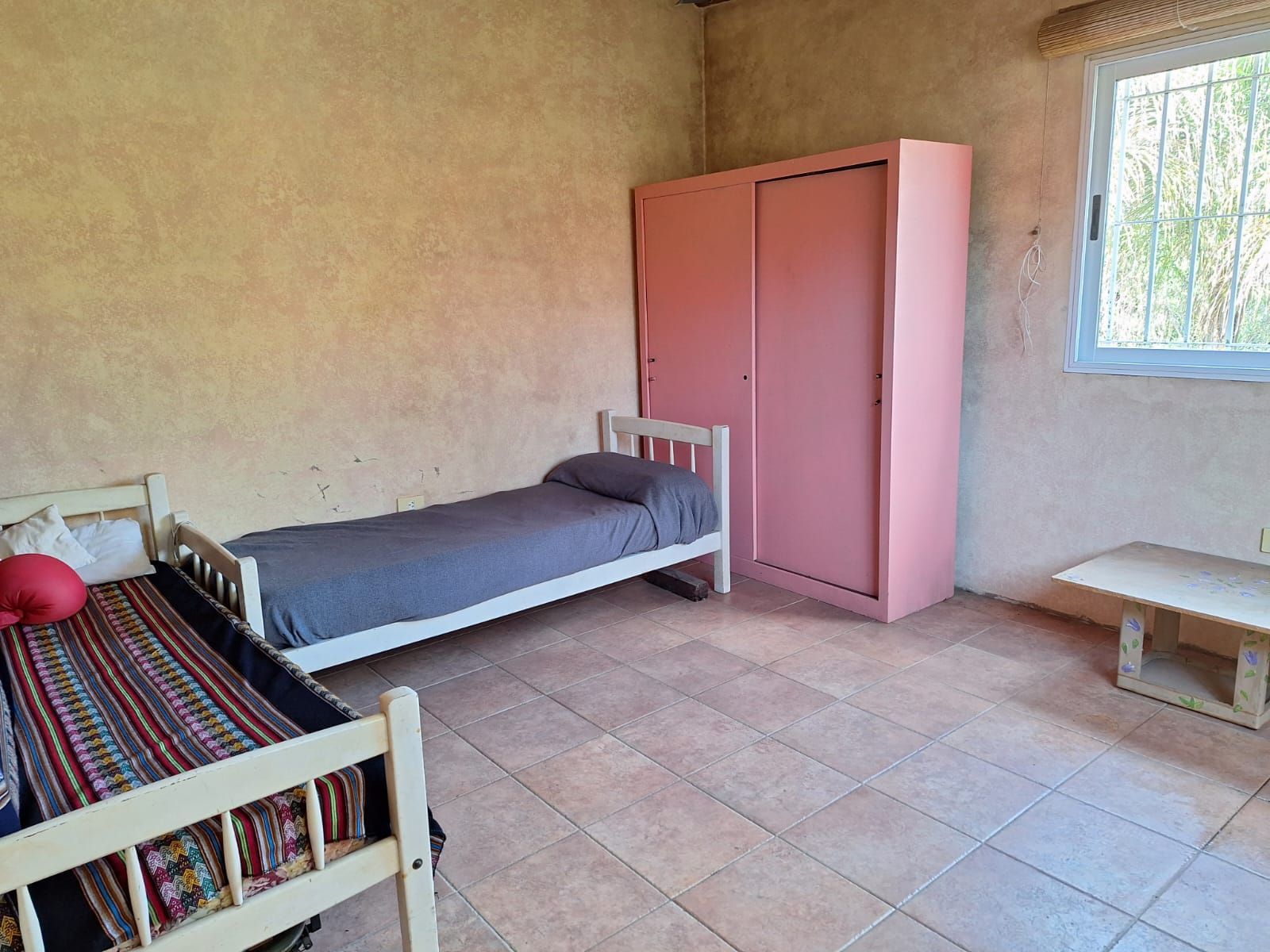 Dormitorio con dos camas individuales, armario rosa, ventana y suelo de baldosas.