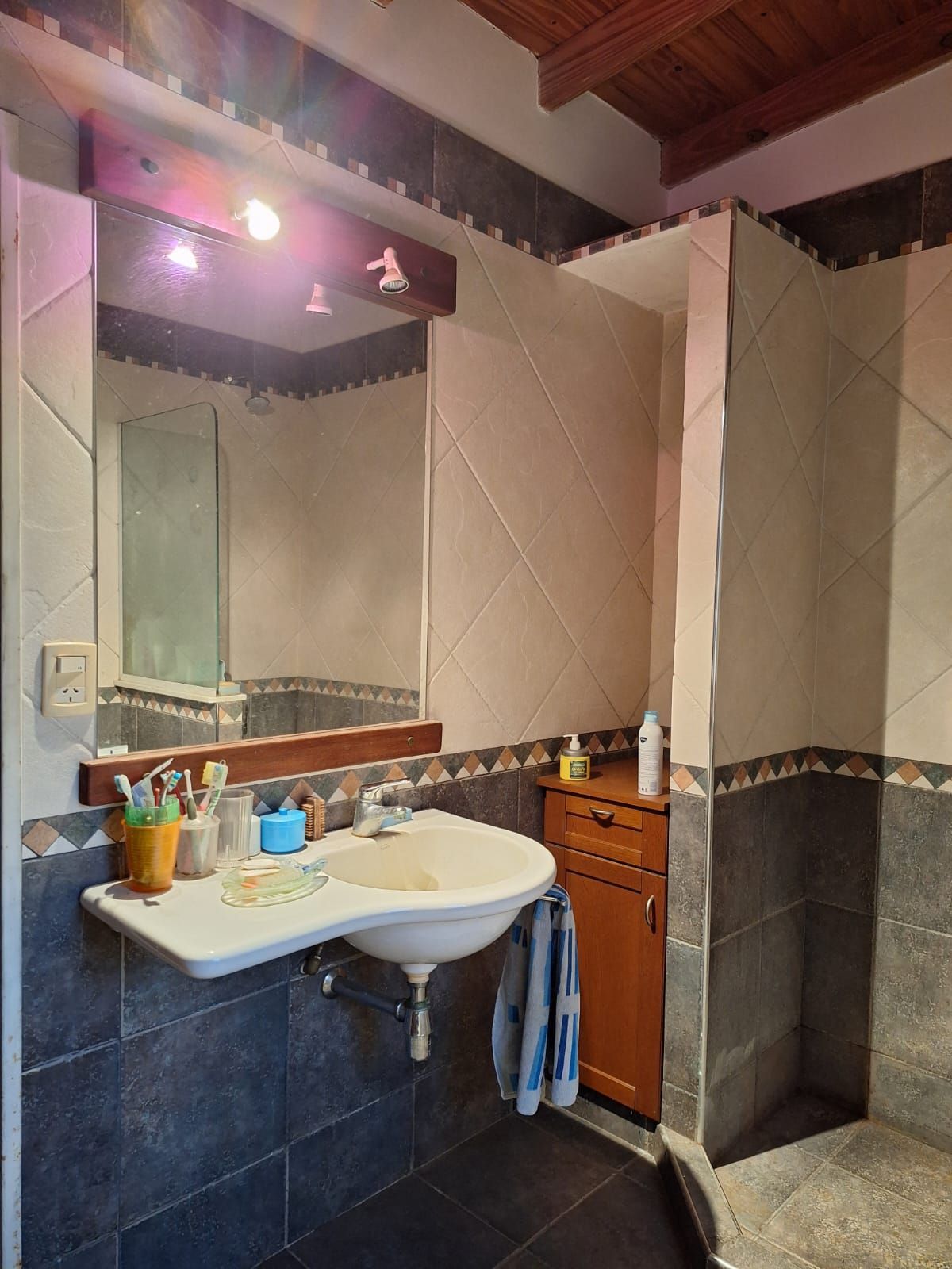 Baño con suelo y paredes de azulejos oscuros, lavabo blanco, armario de madera y espejo grande.