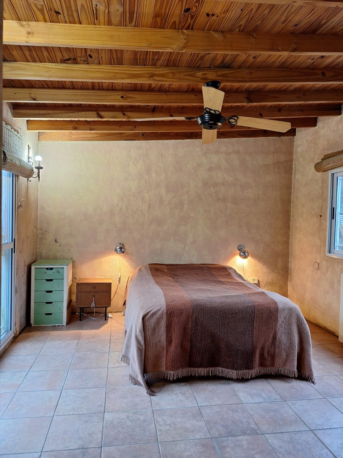 Dormitorio con cama, vigas de madera a la vista en el techo, suelo de baldosas y cómoda pequeña.