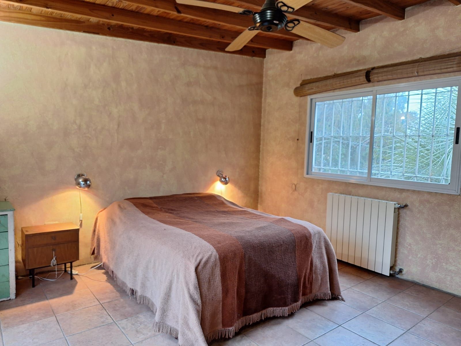 Dormitorio con cama, mesita de noche, ventana, radiador y ventilador de techo. Paredes en tonos tierra y techo de madera.