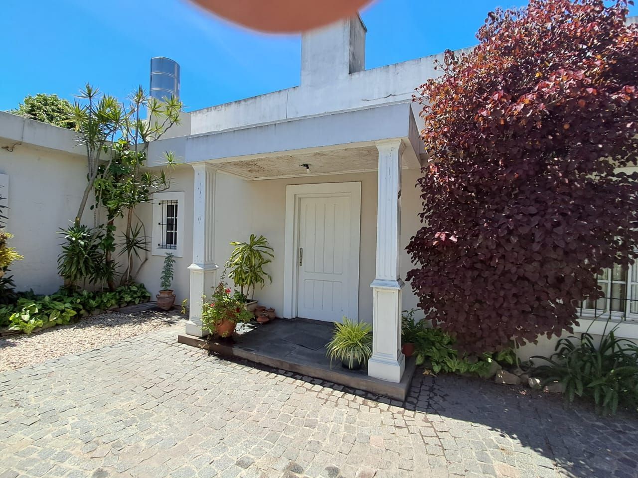 Una casa de color claro con porche y puerta blanca, rodeada de vegetación y con un camino de entrada empedrado.