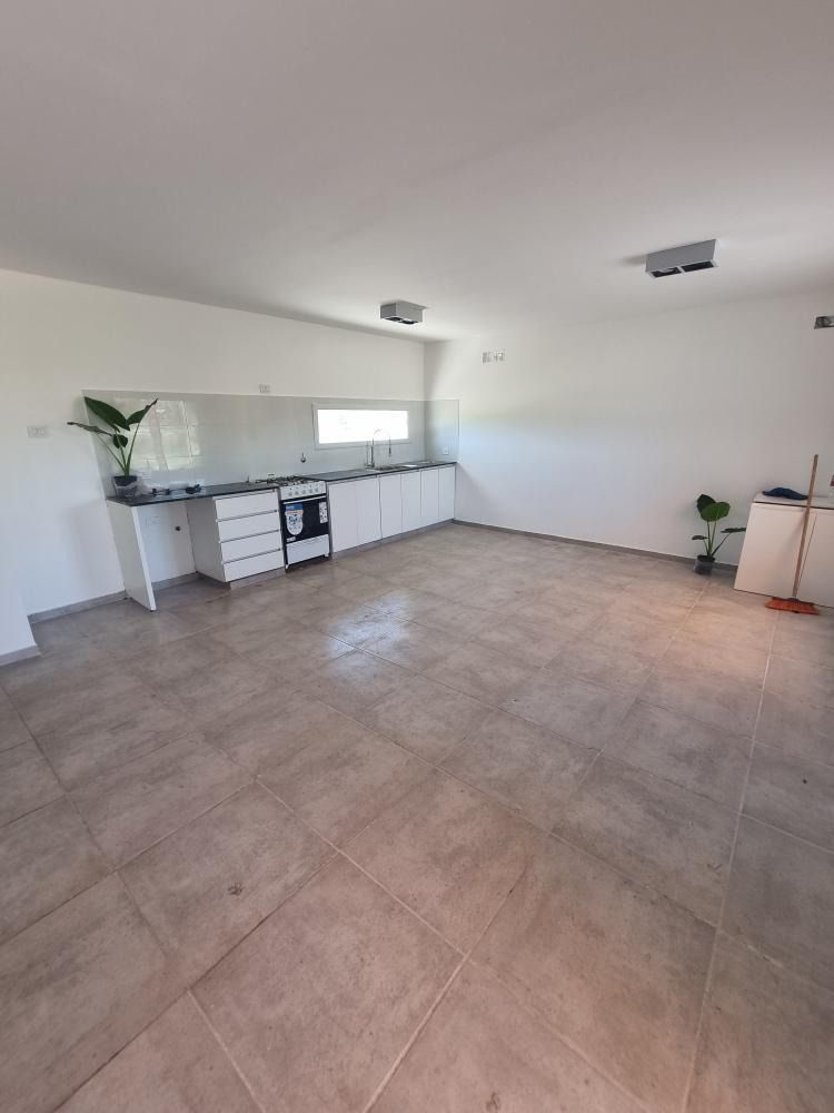 Cocina amplia y vacía con armarios blancos, encimeras oscuras y suelo de baldosas; dos plantas pequeñas.