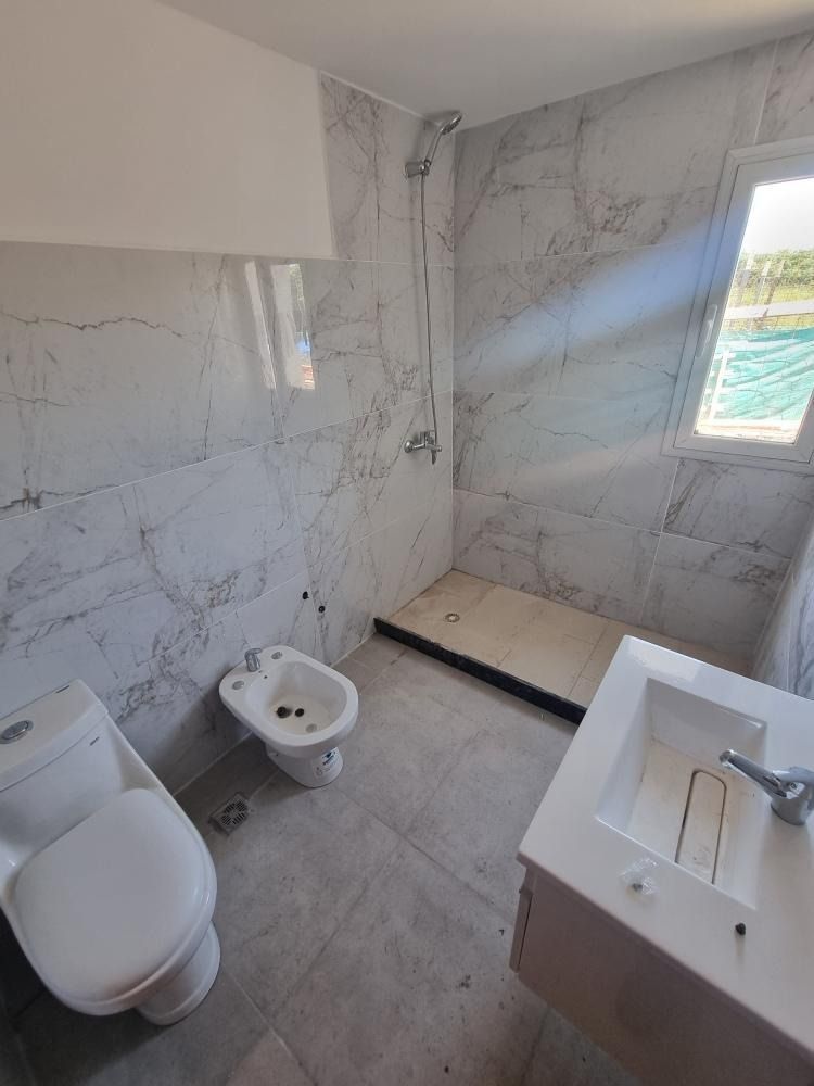 Baño con paredes revestidas con azulejos imitación mármol, inodoro, bidé, ducha y lavabo. Ventana a la derecha.