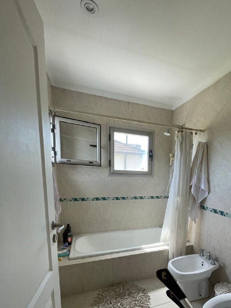 Baño con bañera, ventana e inodoro; azulejos de color claro y cortina.