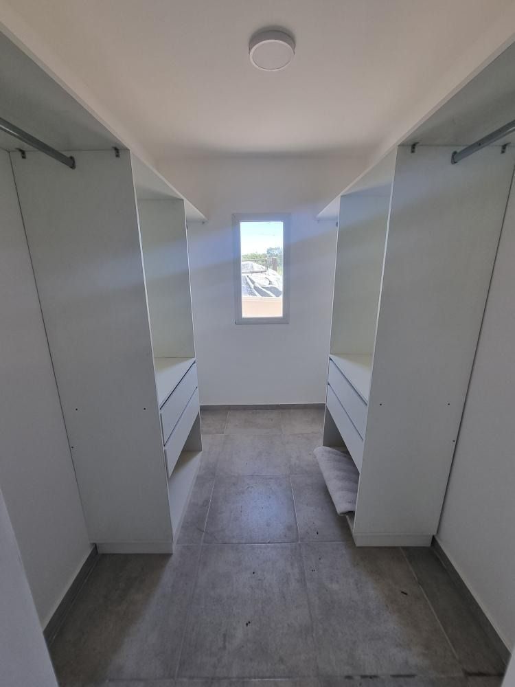Vestidor con estantes y cajones blancos, suelo de cemento y una pequeña ventana al fondo.