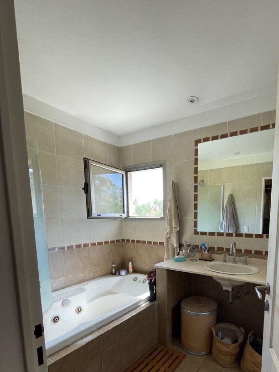 Baño con bañera de hidromasaje, ventana, lavabo y espejo; paredes color crema y detalles de azulejos color canela.