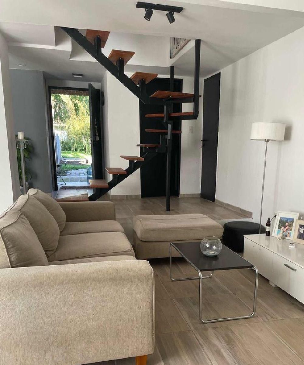 Sala de estar con sofá beige, escalera flotante, mesa de centro y vistas al exterior.