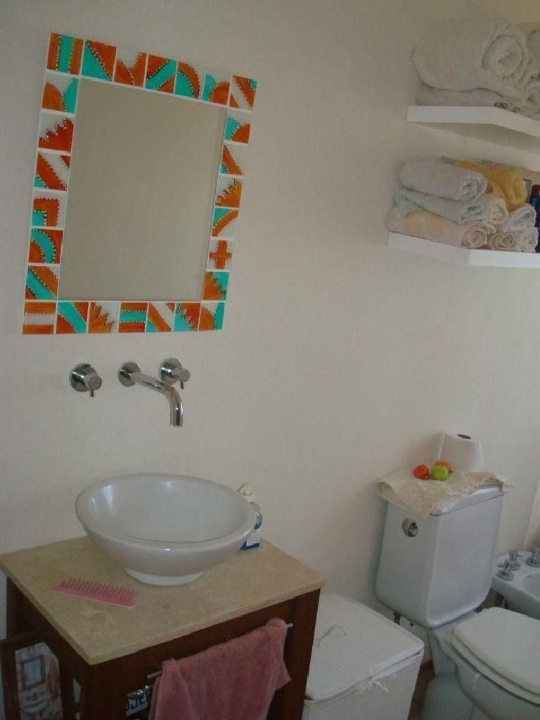 Baño con espejo, lavabo e inodoro. El espejo tiene un marco de mosaico naranja y turquesa. En estantes blancos hay toallas.