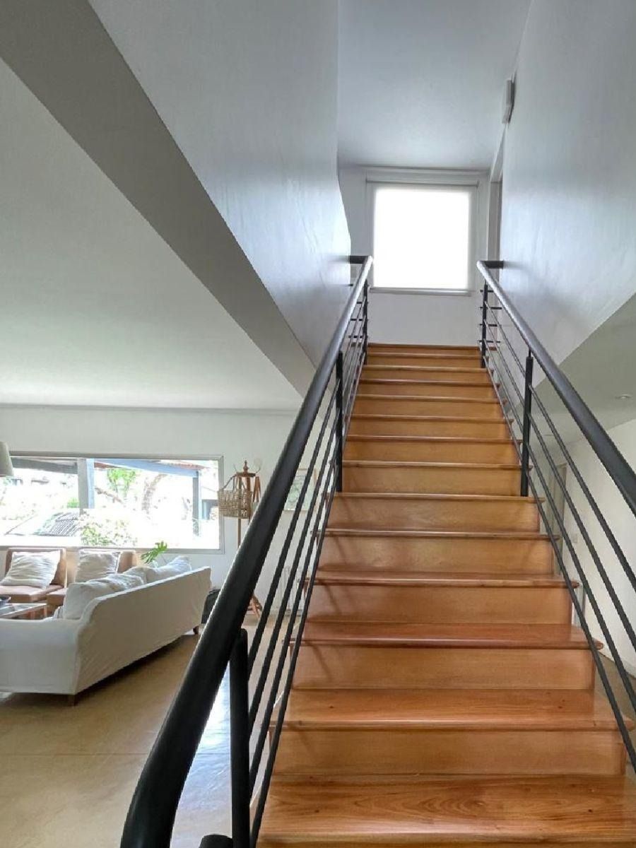 Escalera de madera que conduce hacia arriba, barandillas negras a cada lado, paredes blancas.