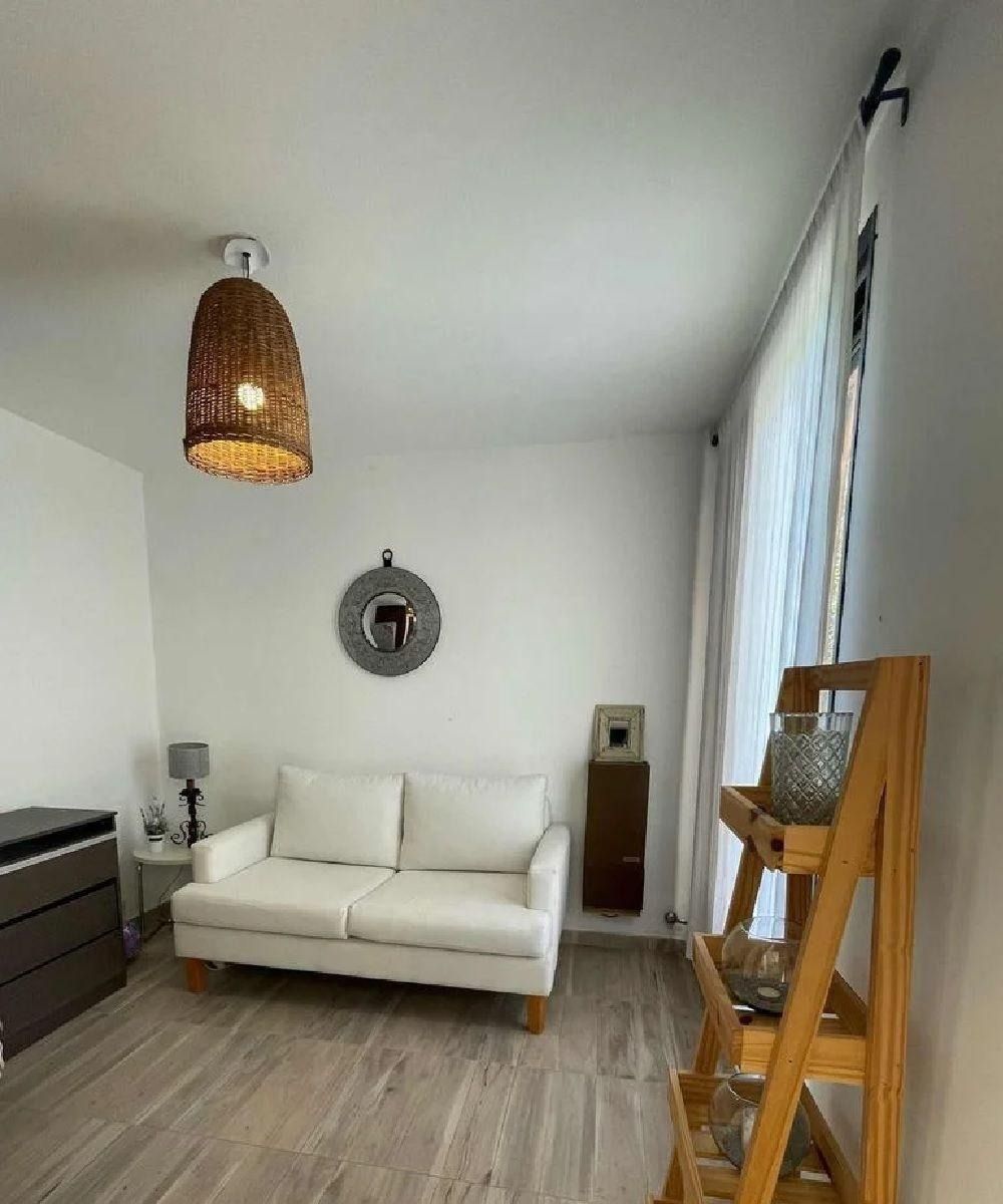 Acogedor salón con paredes blancas, sofá blanco, lámpara de mimbre, espejo decorativo y estanterías de madera.