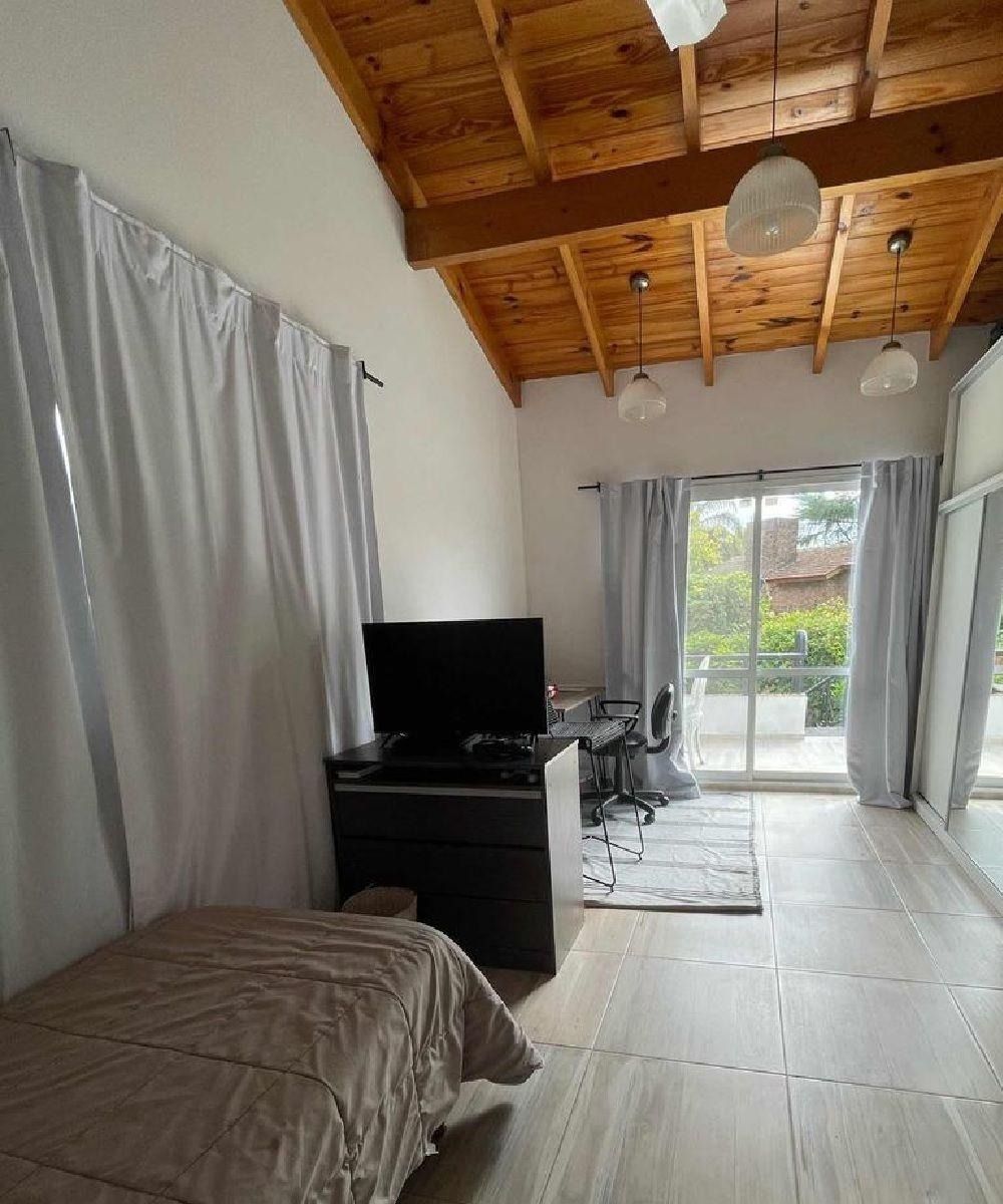 Dormitorio con techo de madera, cama, TV, escritorio y puerta corredera a un balcón.