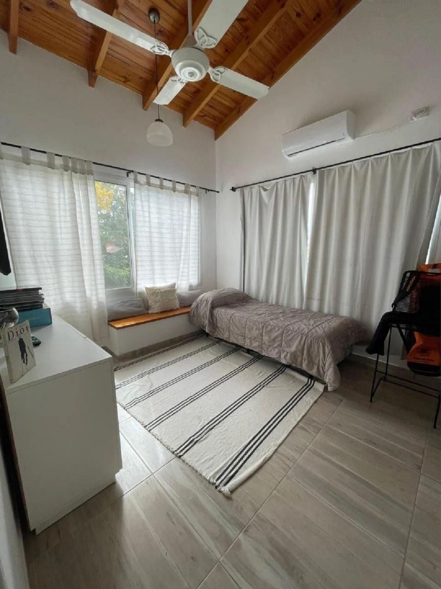 Dormitorio con paredes blancas, techo de madera y cama con una manta marrón claro. Cortinas blancas y alfombra de rayas.