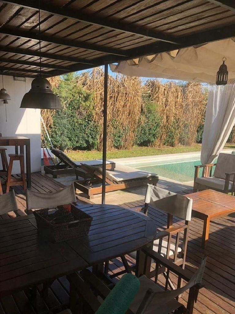 Patio exterior con muebles de madera, piscina y plantas. Zona de estar cubierta.