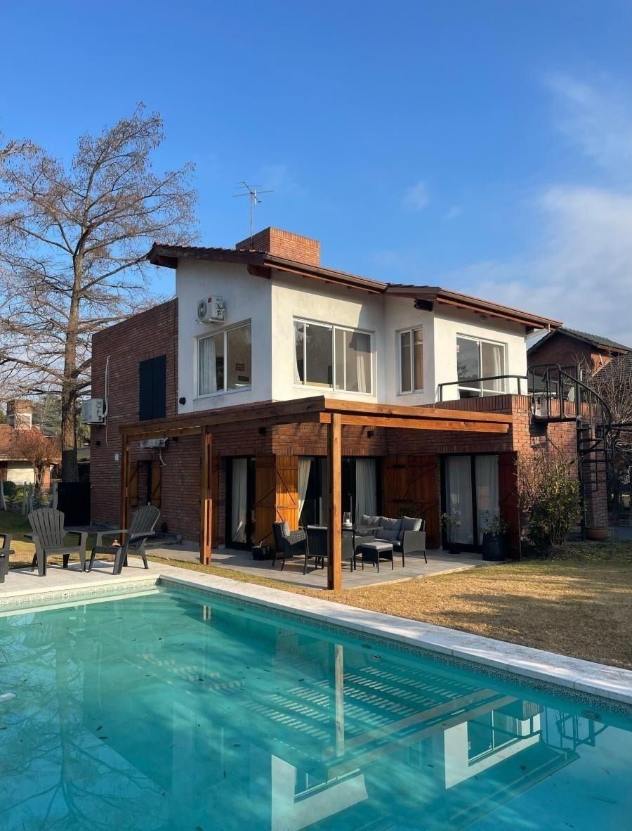Casa de dos plantas con piscina. Exterior blanco y ladrillo, pérgola de madera y cielo azul.