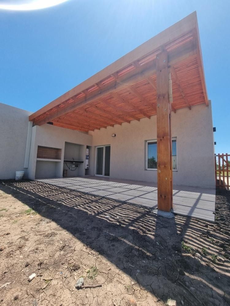 Pérgola de madera que cubre un patio junto a una casa con parrilla empotrada, en un día soleado.