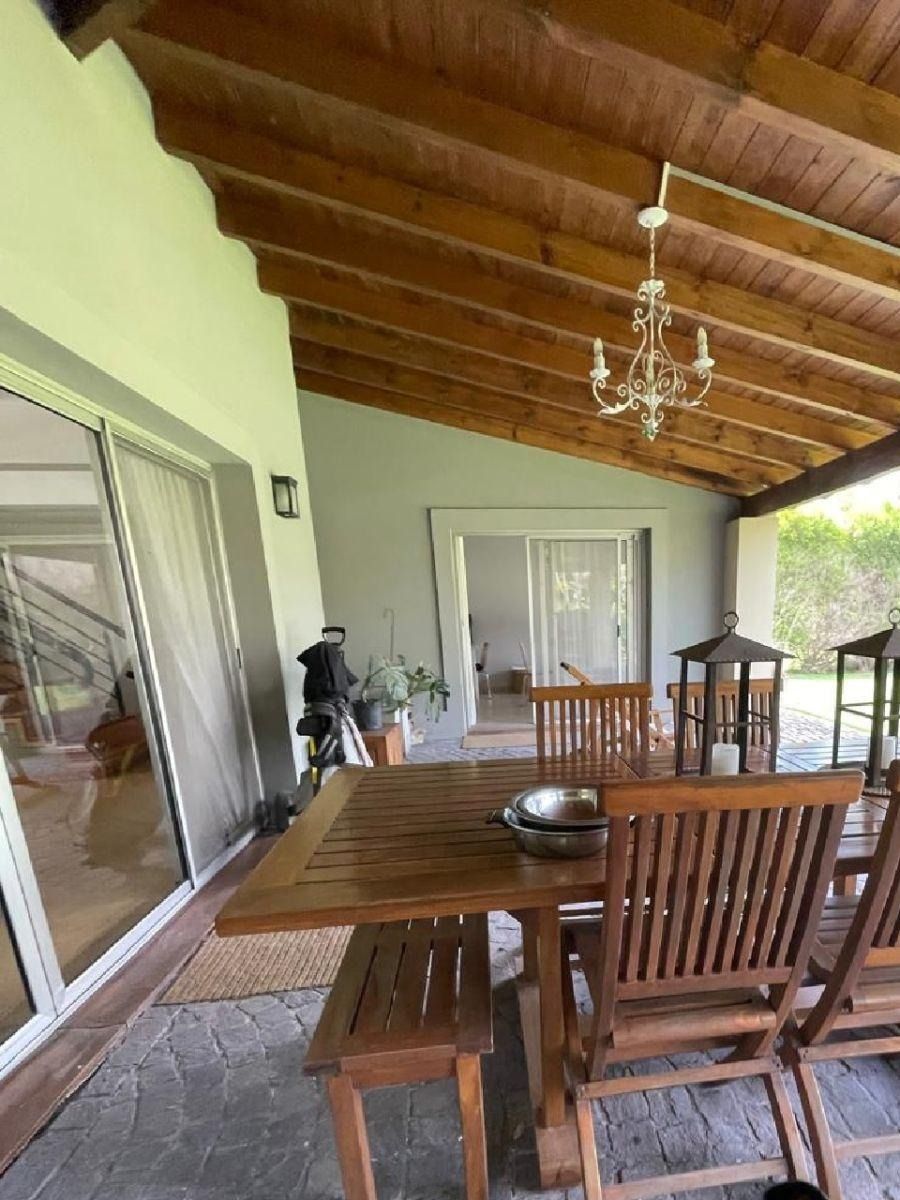 Comedor al aire libre con mesa, sillas y banco de madera bajo un patio cubierto con lámpara de araña.