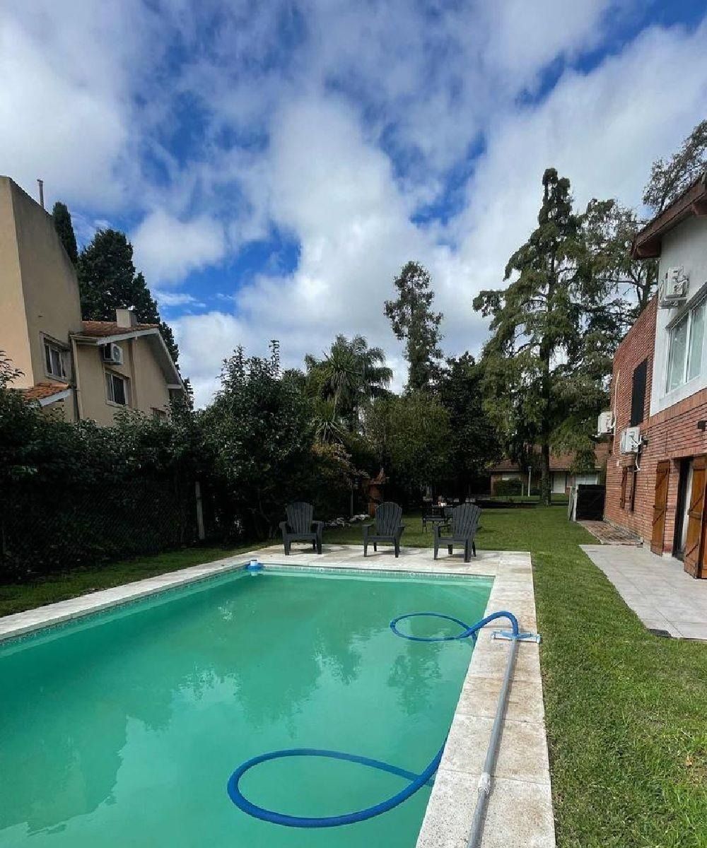 Piscina en el patio trasero de edificios de dos plantas, cielo azul. Hay césped, árboles y tumbonas.