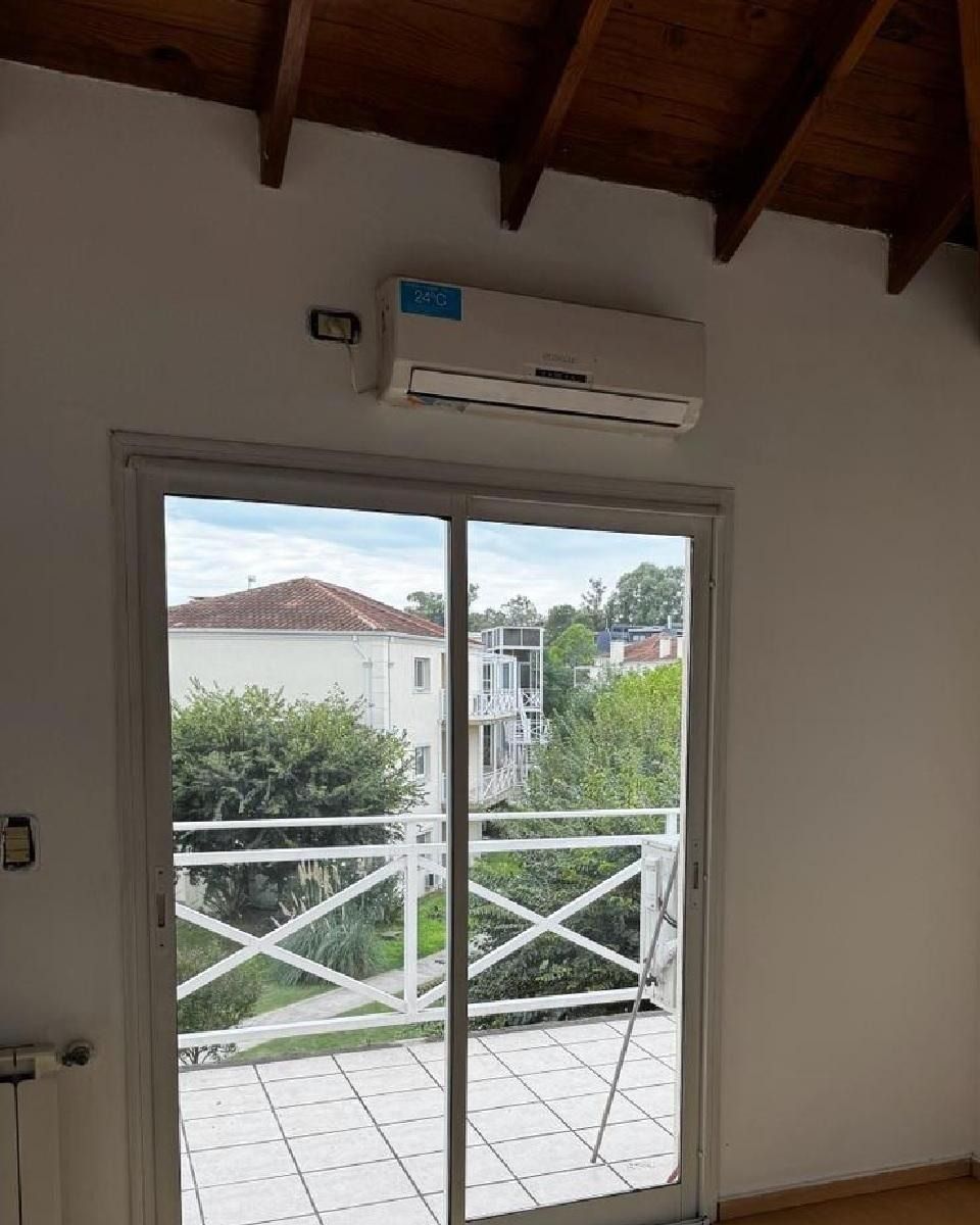 Aire acondicionado sobre una puerta de cristal que da a un balcón con barandilla blanca, con vistas a edificios y árboles.