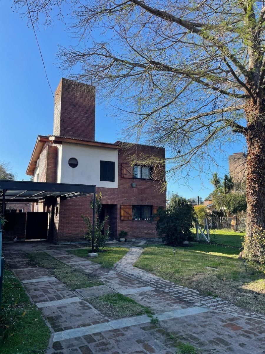 Casa de ladrillo con chimenea, entrada para coches, césped y árbol bajo un cielo azul.