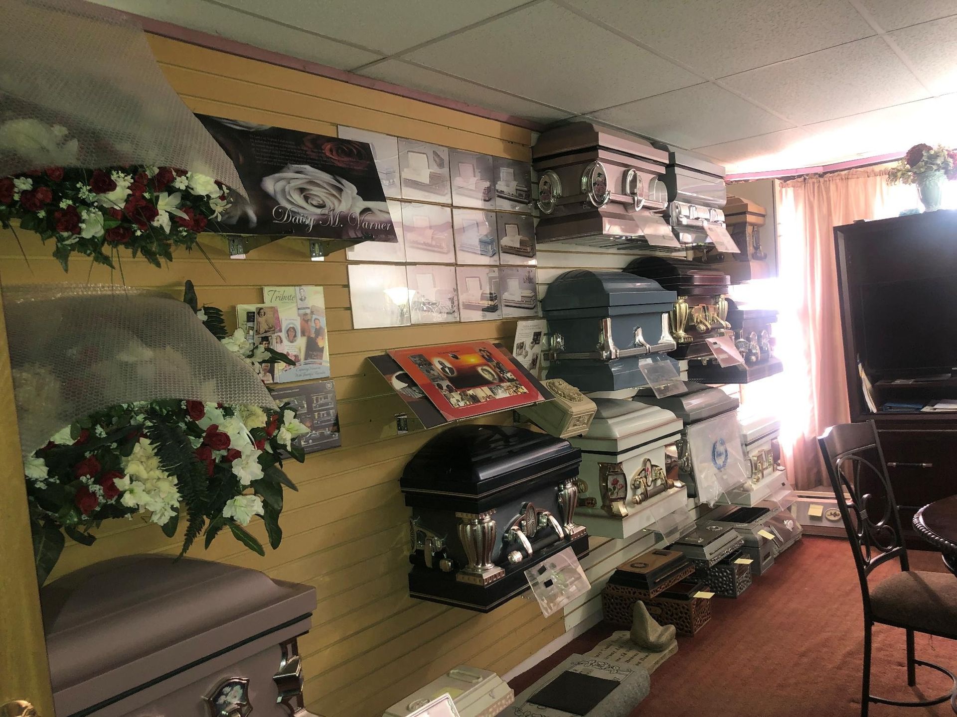 Interior of a funeral home, display of coffins on shelves against a wall, with a door and window visible.