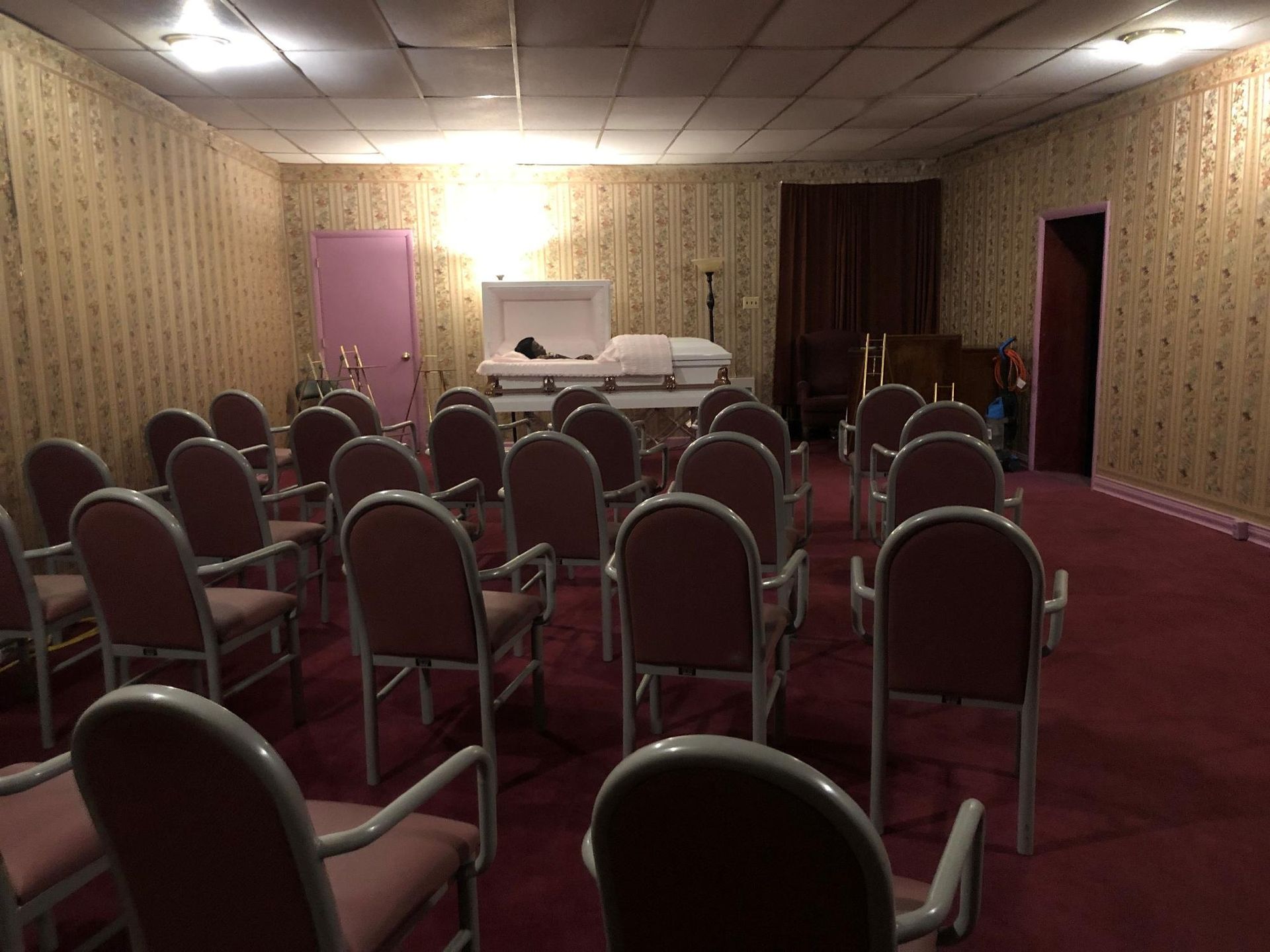 Funeral home chapel with casket, rows of chairs, and patterned wallpaper.